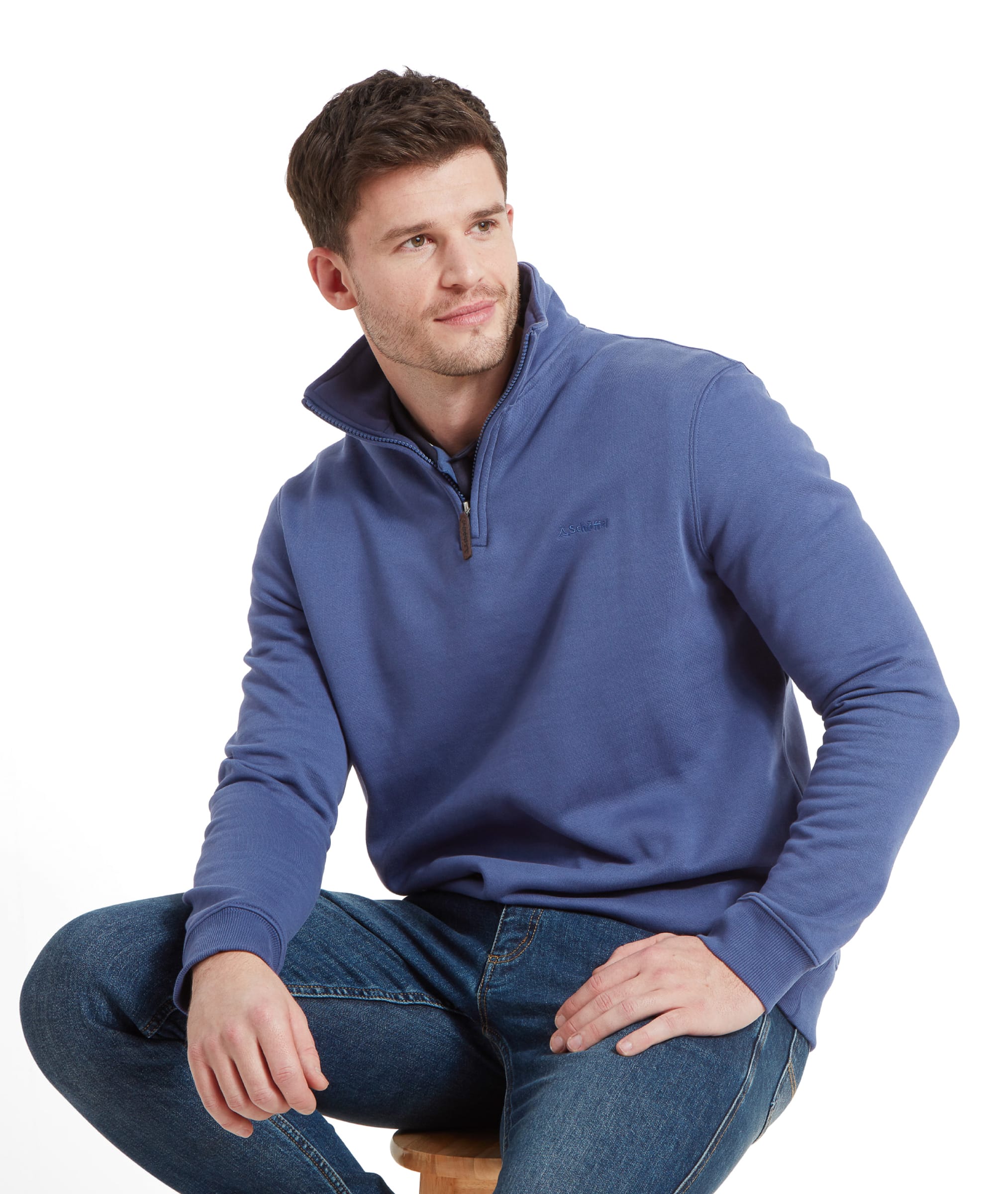 A young man sits casually on a stool, wearing a Schöffel St Merryn Sweatshirt for Men in Light Blue. The sweatshirt features a minimalistic design with a small embroidered brand logo on the left chest. He pairs it with blue jeans and has short brown hair, giving a relaxed, approachable appearance.