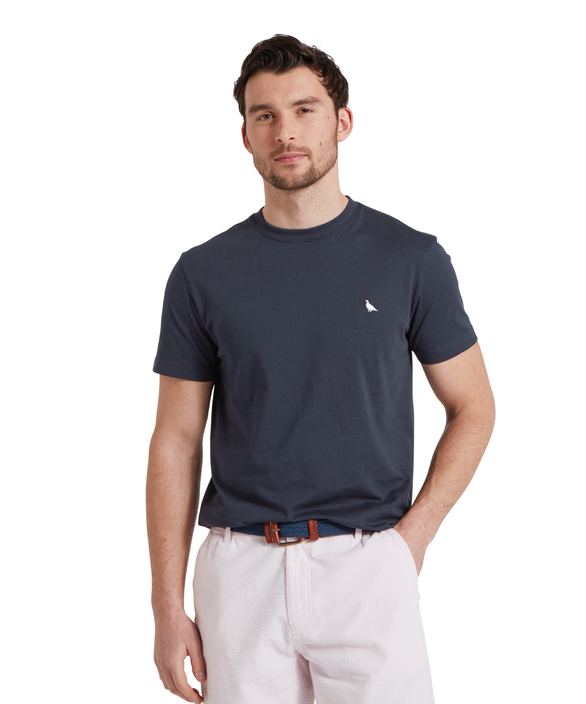 Close-up of a male model wearing a navy Schöffel t-shirt with a small embroidered white bird logo on the chest, paired with light grey shorts and a blue woven belt. He is standing in a relaxed pose with one hand his pocket.