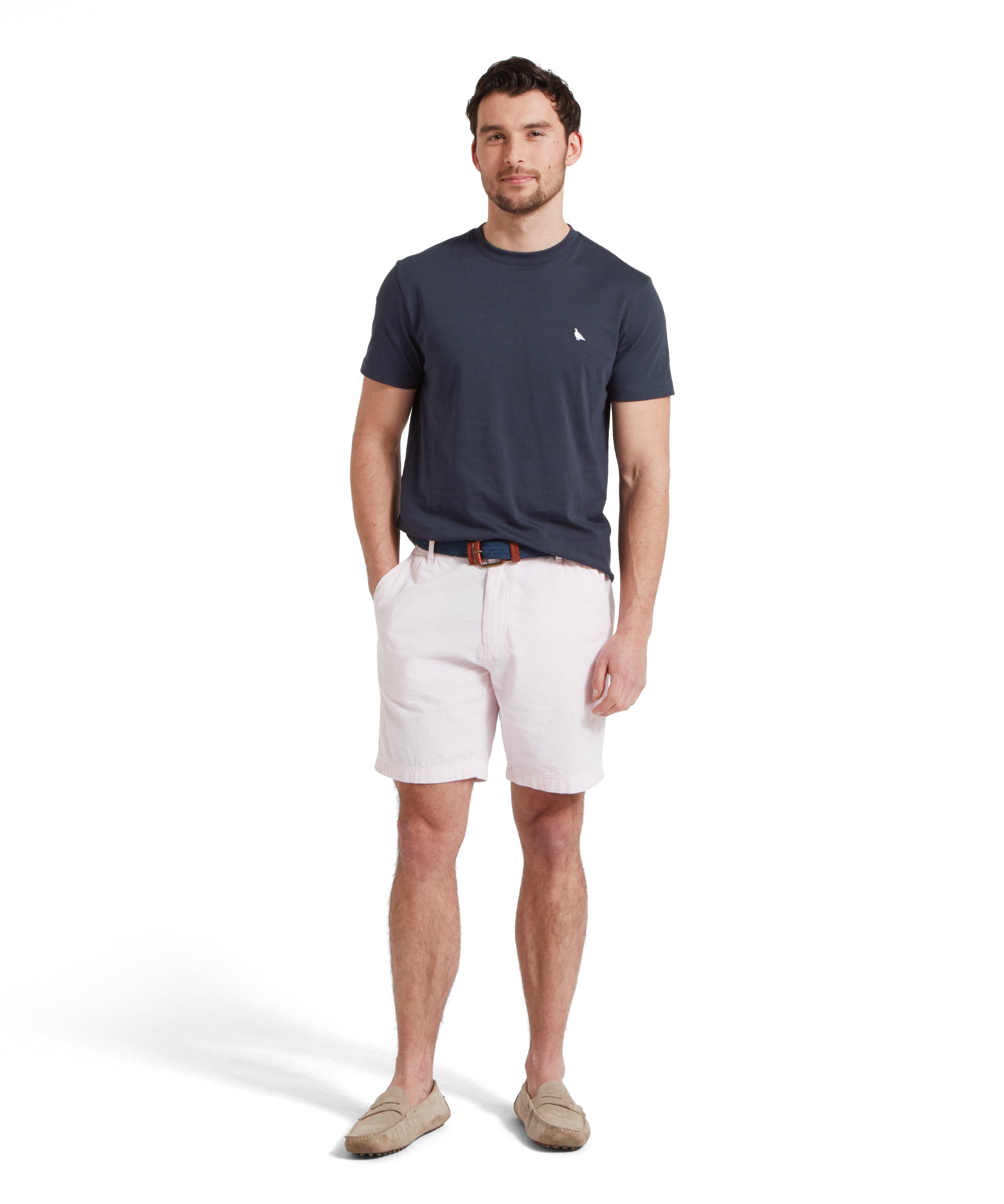 Full-height image of a male model wearing a navy Schöffel t-shirt with a small embroidered white bird logo on the chest, paired with light grey shorts, tan loafers and a blue woven belt. He is standing in a relaxed pose with one hand his pocket.