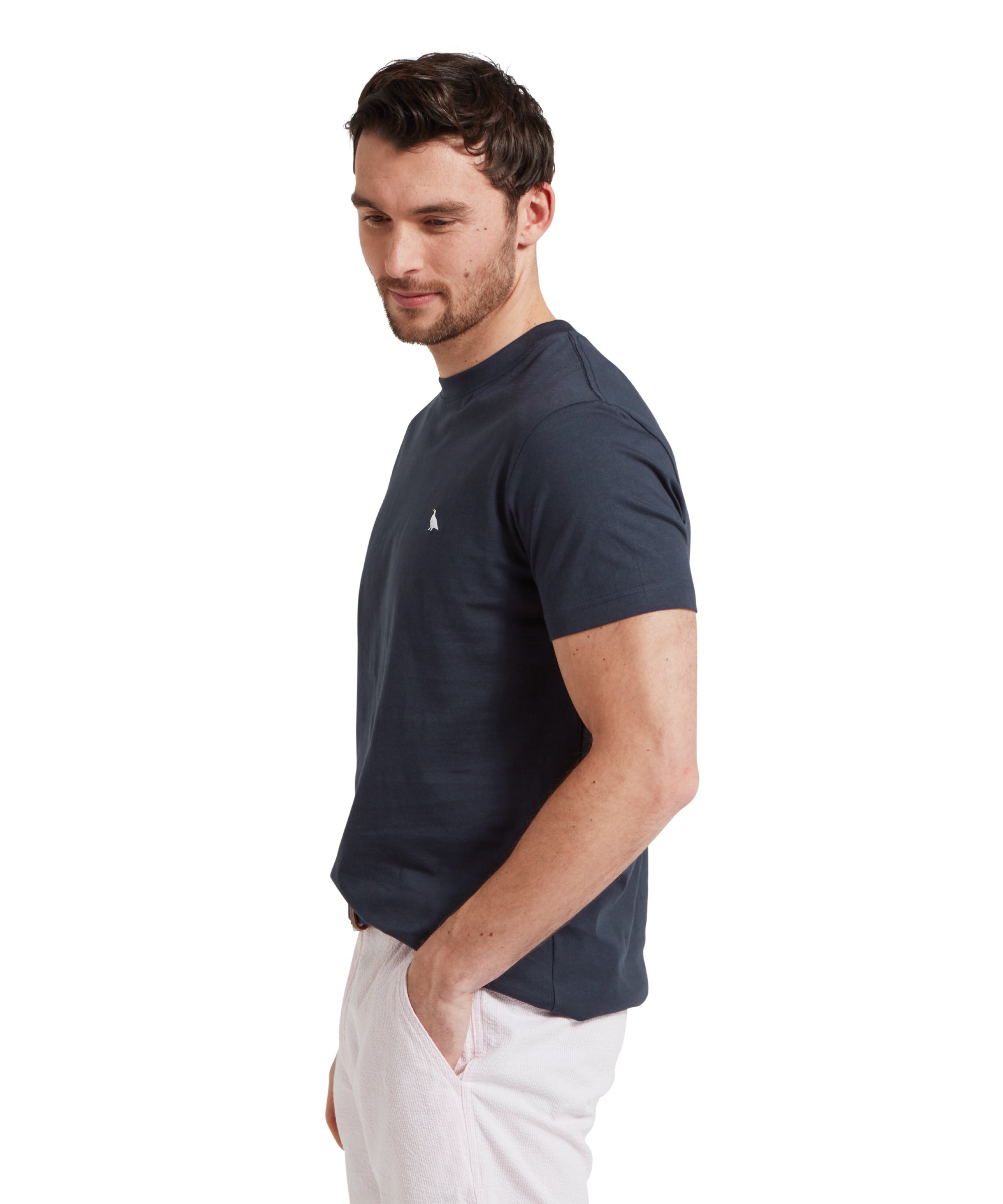 Side profile of a male model wearing a navy Schöffel t-shirt with a small embroidered white bird logo on the chest, paired with light grey shorts and a blue woven belt. He is standing in a relaxed pose with one hand his pocket.