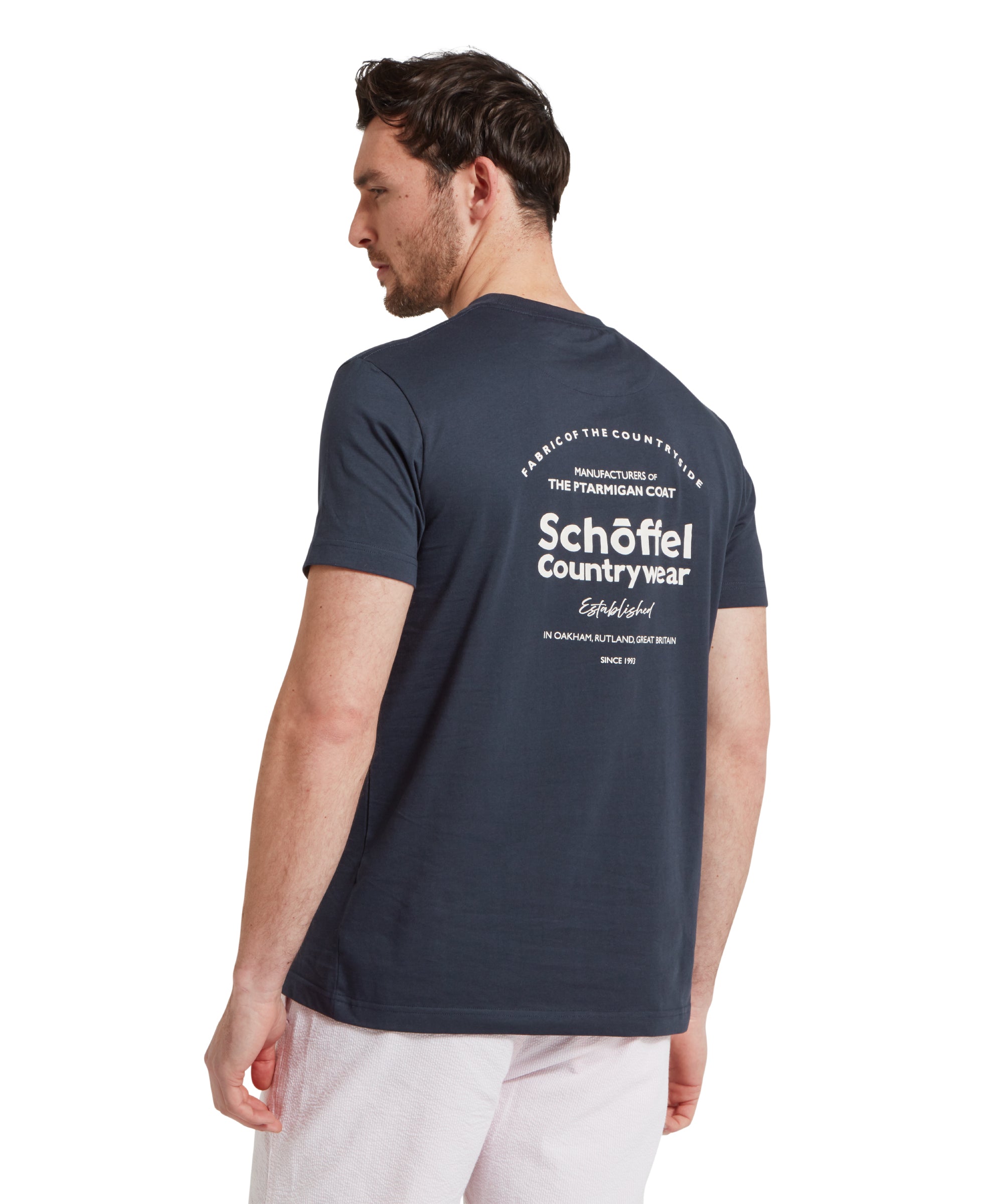 Back image of a male model wearing a navy Schöffel t-shirt with a large "Schöffel Countywear" white logo on the back, paired with light grey shorts and a blue woven belt. He is standing in a relaxed pose with his hands by his side.