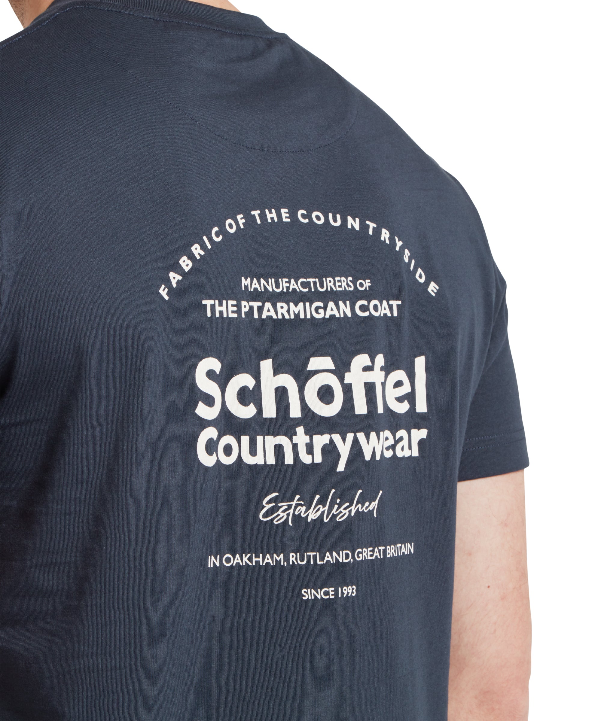 Close up of a male model wearing a navy Schöffel t-shirt with a large "Schöffel Countywear" white logo on the back. The image highlights the white logo.