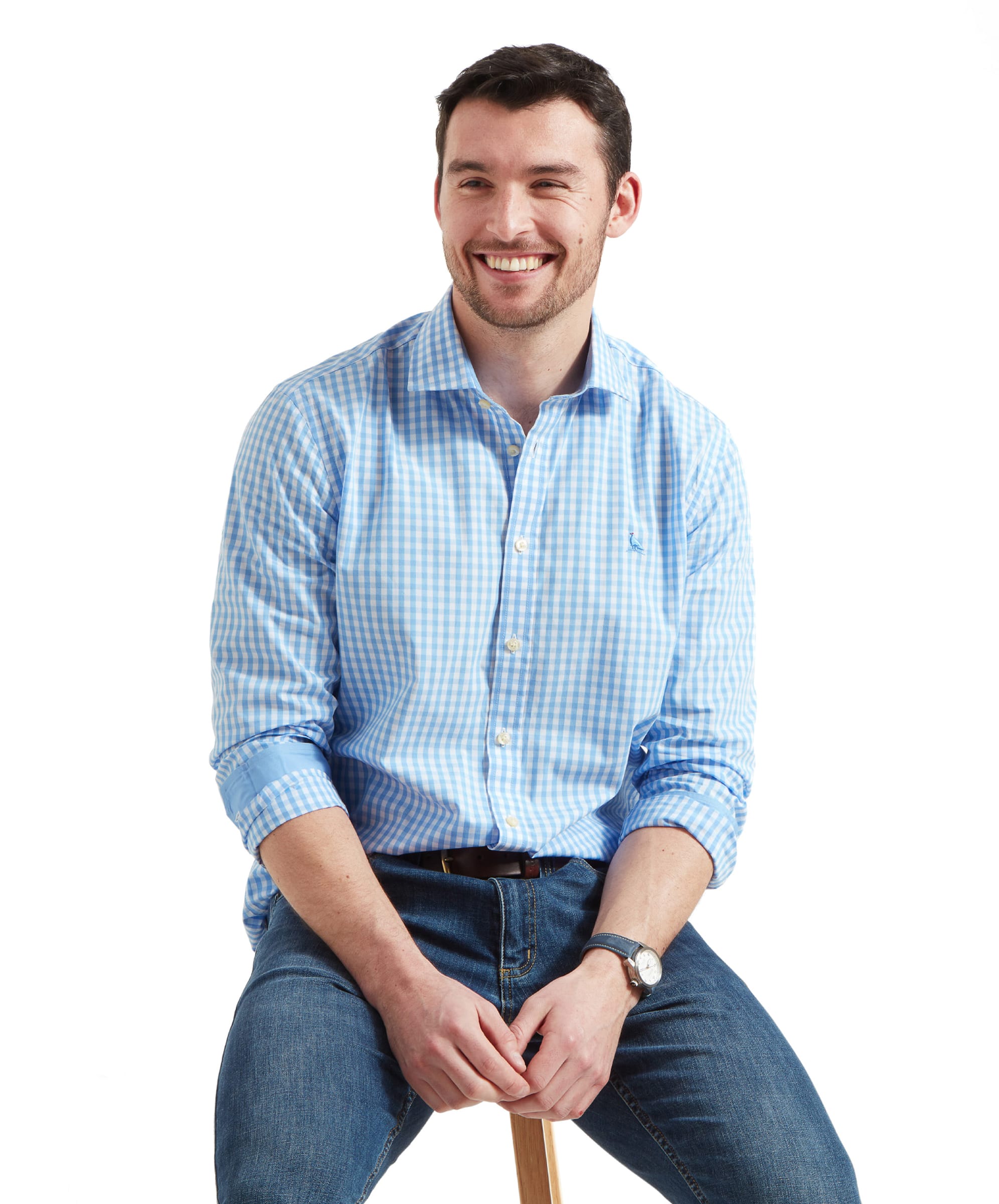 A man is sitting on a stool, wearing a Schöffel Thorpeness Tailored Shirt for Men in Blue with rolled-up sleeves, paired with jeans. He is smiling and looking to the side, showcasing the shirt's pattern and relaxed fit.