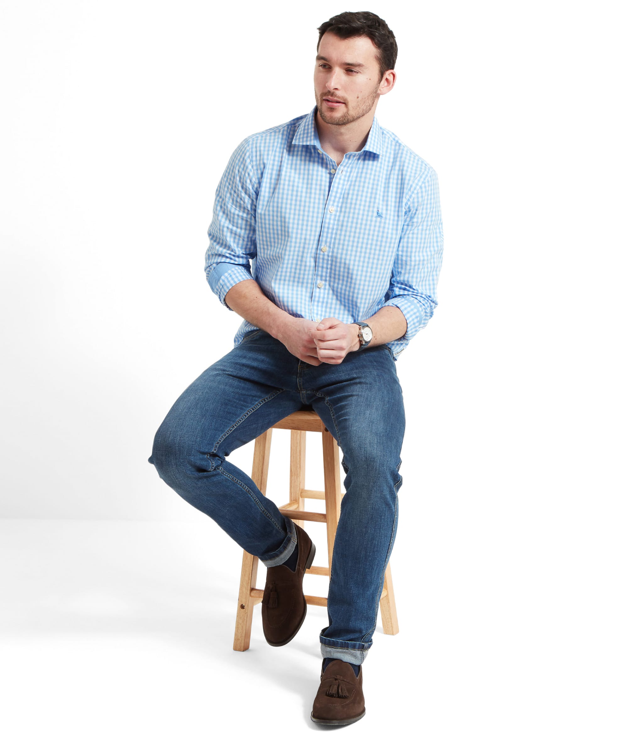 The same man is seated, slightly leaning forward. The outfit includes the Schöffel Thorpeness Tailored Shirt for Men in Blue, jeans, and brown suede loafers. His watch and belt add to the smart-casual look.