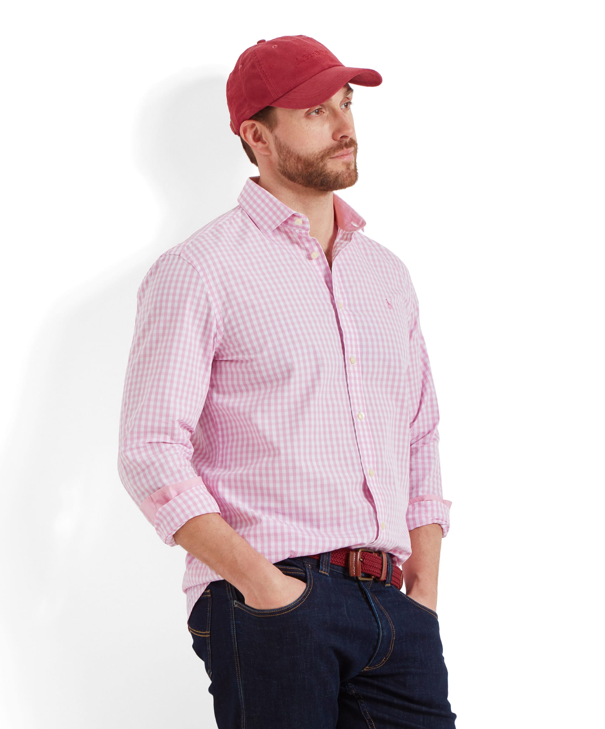 A man is wearing a Schöffel Thorpeness Tailored Shirt for Men in Pink with the sleeves rolled up and a red cap. He is standing with his hands in his pockets, looking to the side. The shirt is paired with dark jeans.