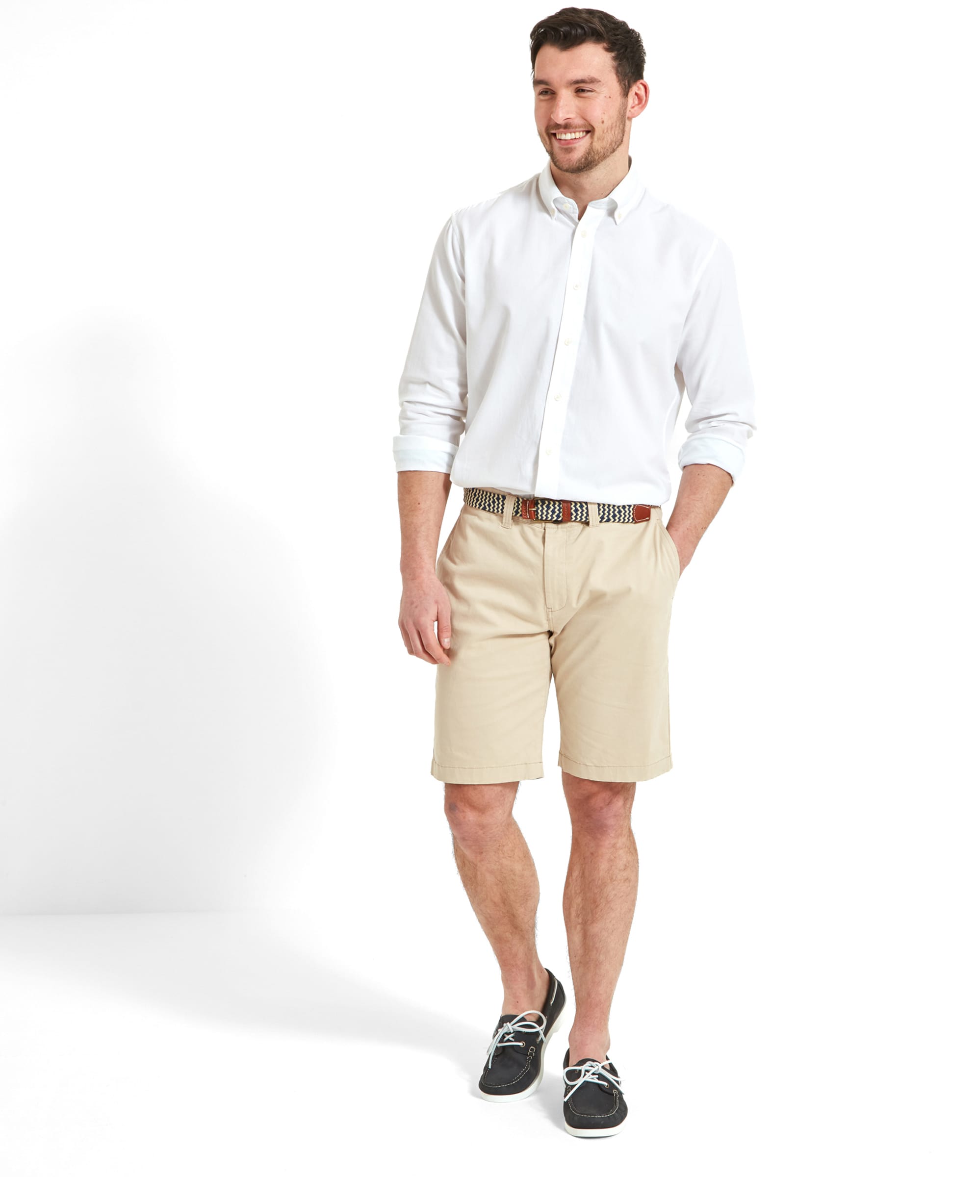 The man is dressed in a Schöffel Titchwell Tailored Shirt for Men in White paired with beige shorts. He is smiling broadly, giving off a friendly and relaxed vibe. His outfit is completed with a woven belt and dark boat shoes, ideal for a casual, warm-weather look.