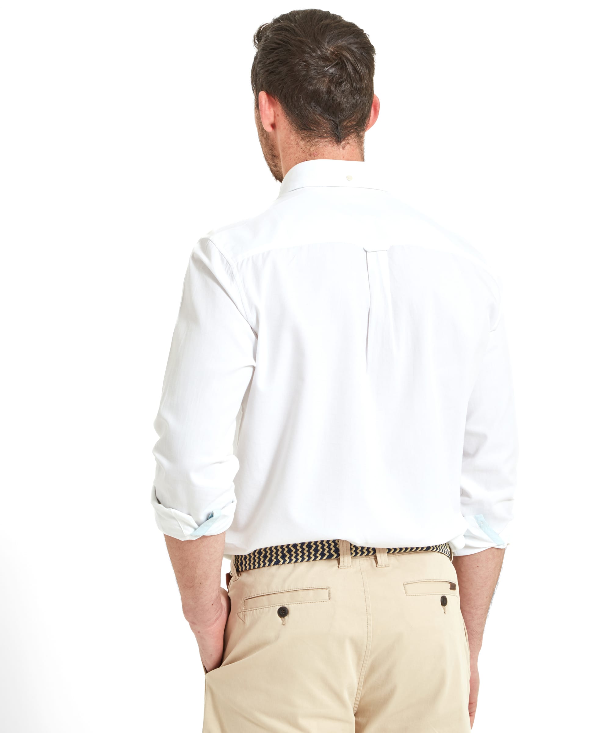 The man is seen from the back, showcasing the fit and style of his Schöffel Titchwell Tailored Shirt for Men in White. The shirt has a classic design with a pleat at the back. He pairs it with beige shorts, secured with a woven belt, and his hands are casually in his pockets.