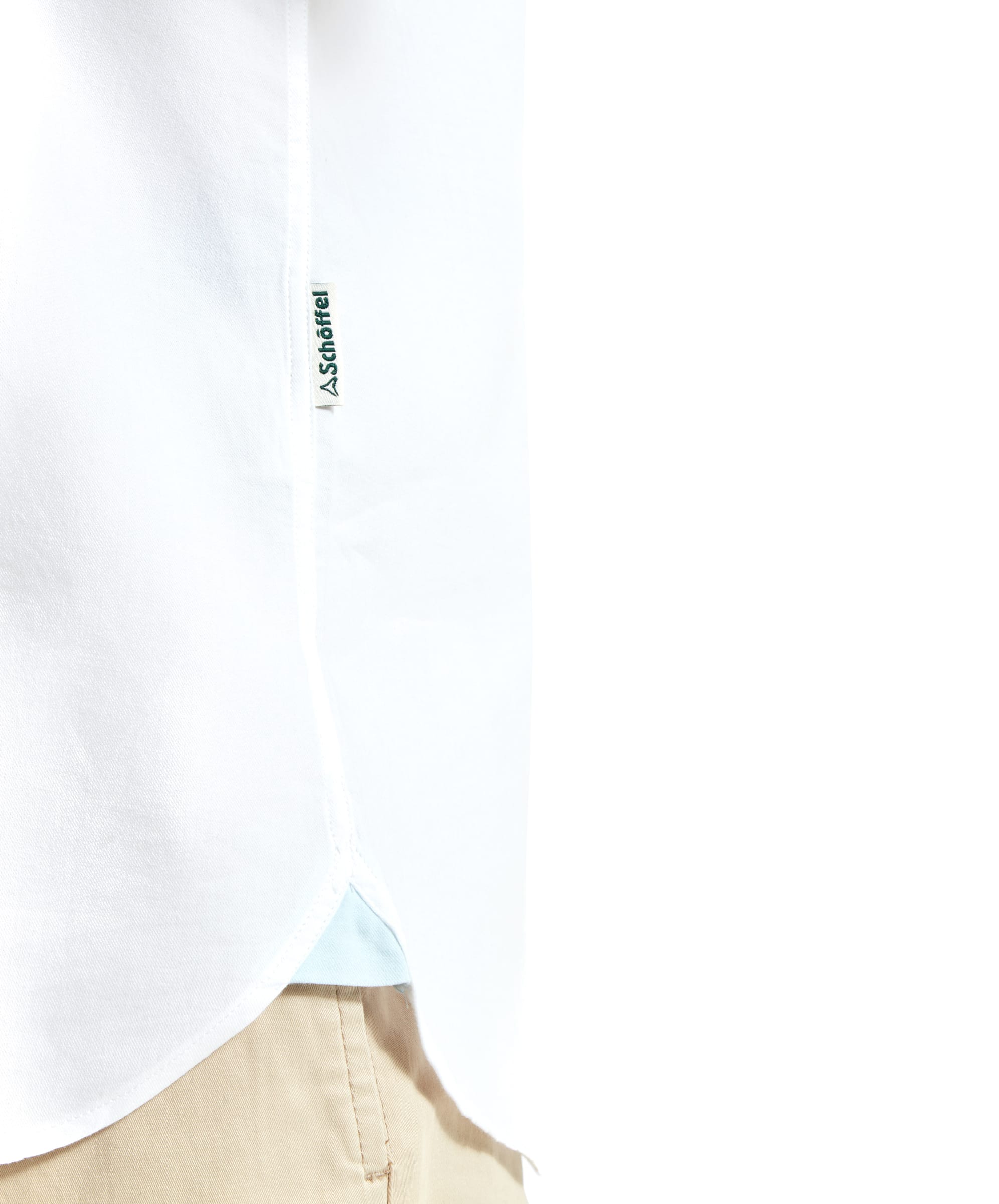 The image focuses on the hem of the Schöffel Titchwell Tailored Shirt for Men in White, highlighting a small brand label sewn into the side seam. The shirt is tucked into beige shorts, giving a clear view of the neat, tailored finish.