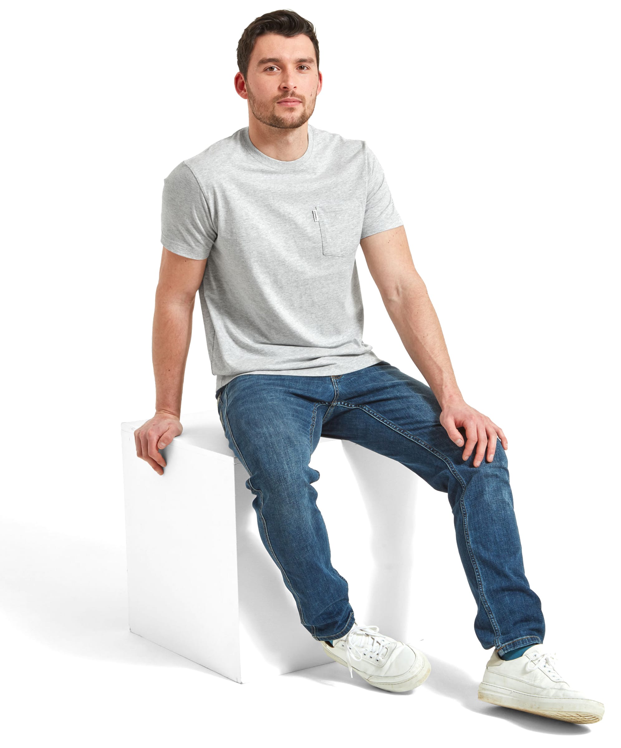 The man is sitting on a white block, wearing the same Schöffel Towan T-Shirt for Men in Grey and jeans, paired with white trainers. His expression is neutral, and he is looking slightly to the side.