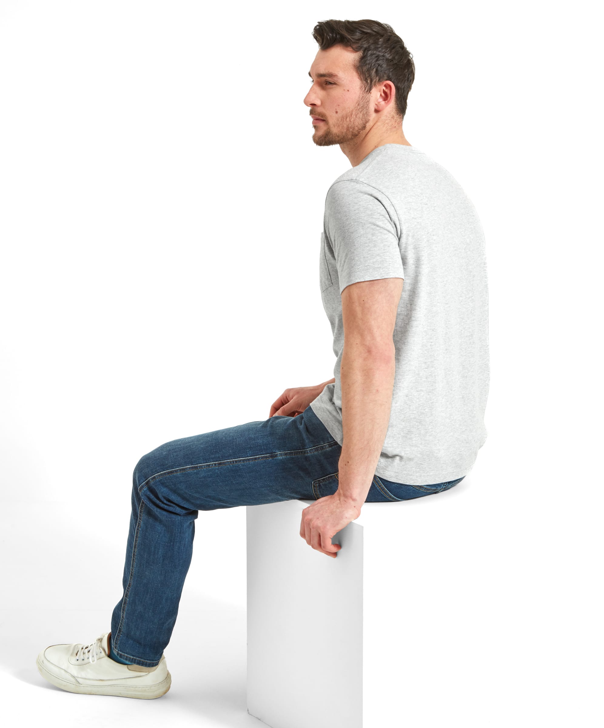 A side view of the man sitting on the white block, still in the Schöffel Towan T-Shirt for Men in Grey and jeans. He is looking off into the distance, giving a profile view.