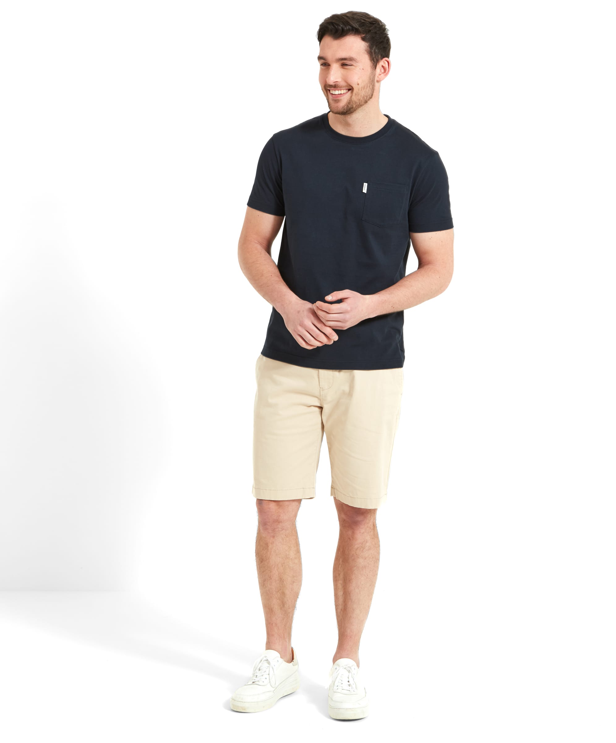 A man stands smiling, wearing a Schöffel Towan T-Shirt for Men in Navy with a small chest pocket and a brand tag. He pairs the T-shirt with beige shorts and white trainers, presenting a casual and comfortable summer look.