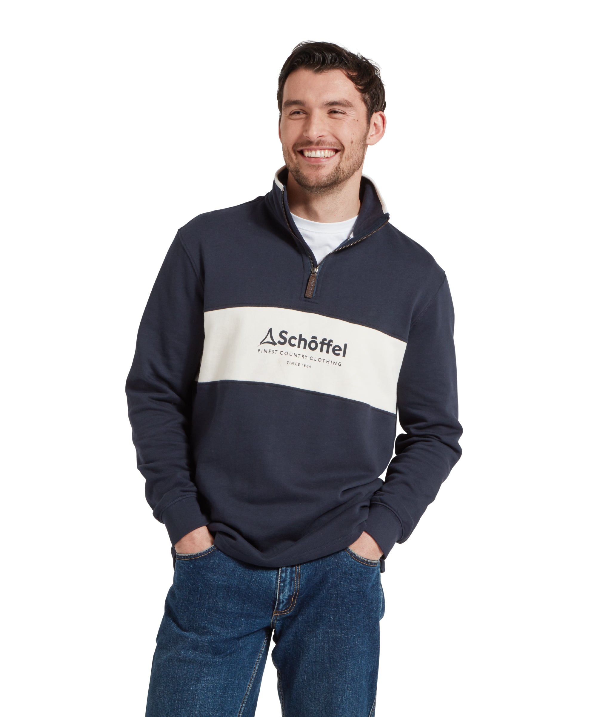 Half height image of a male model wearing a blue Schöffel sweatshirt with a white stripe with blue large embroidered Schöffel logo across the chest. It is paired with a white t-shirt and dark blue jeans and he is standing in a relaxed pose with his hands in his pockets and he is smiling looking away from the camera.