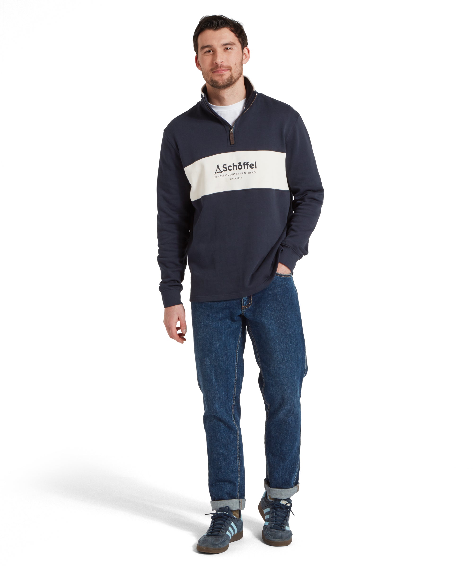 Full height image of a male model wearing a blue Schöffel sweatshirt with a white stripe with blue large embroidered Schöffel logo across the chest. It is paired with a white t-shirt and dark blue jeans and he is standing in a relaxed pose one hand in his pocket and he is smiling looking towards the camera.