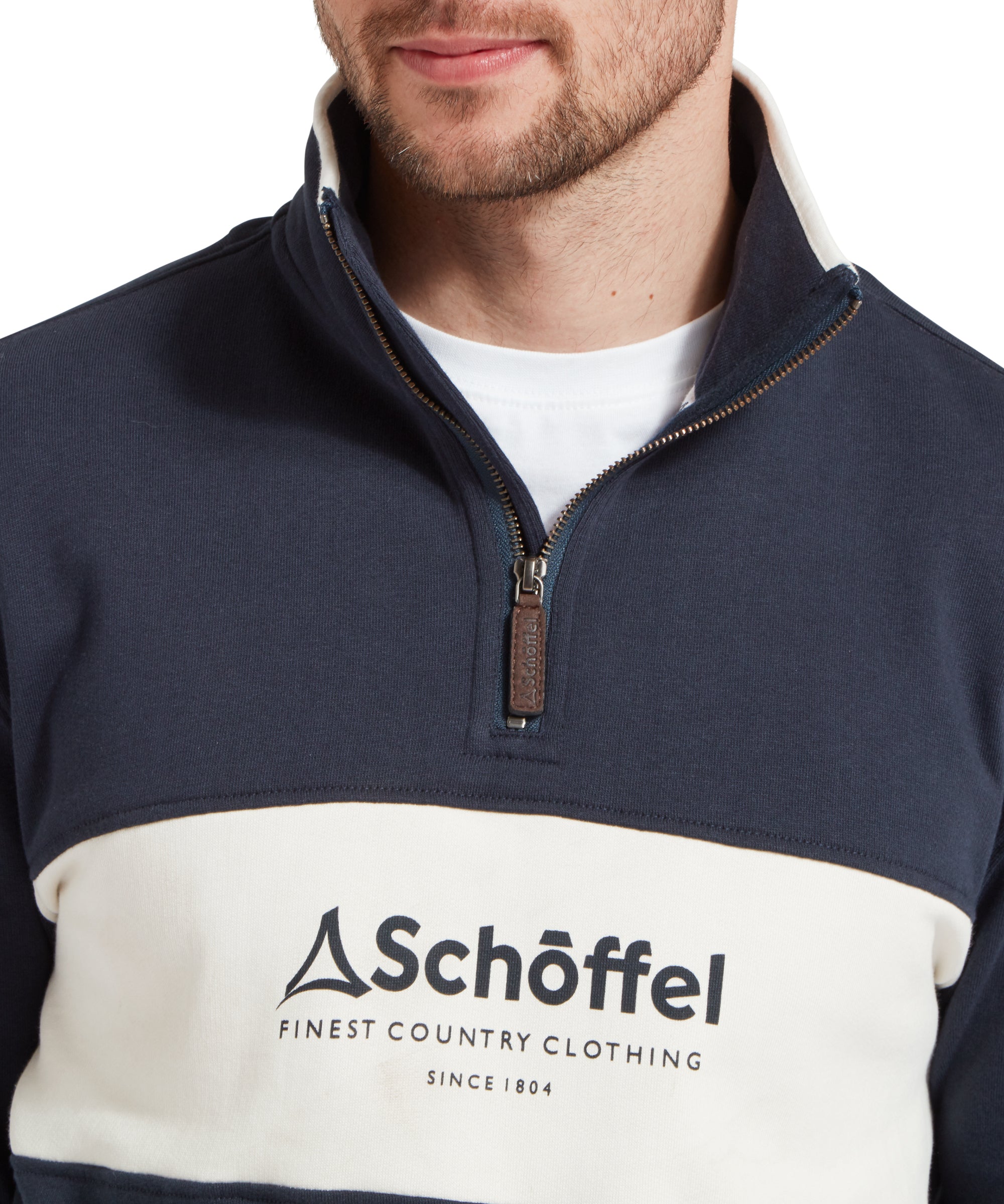 Close up image of a male model wearing a blue Schöffel sweatshirt with a white stripe with blue large embroidered Schöffel logo across the chest. It is paired with a white t-shirt and dark blue jeans. Image highlights quarter zip neck with pull tab.