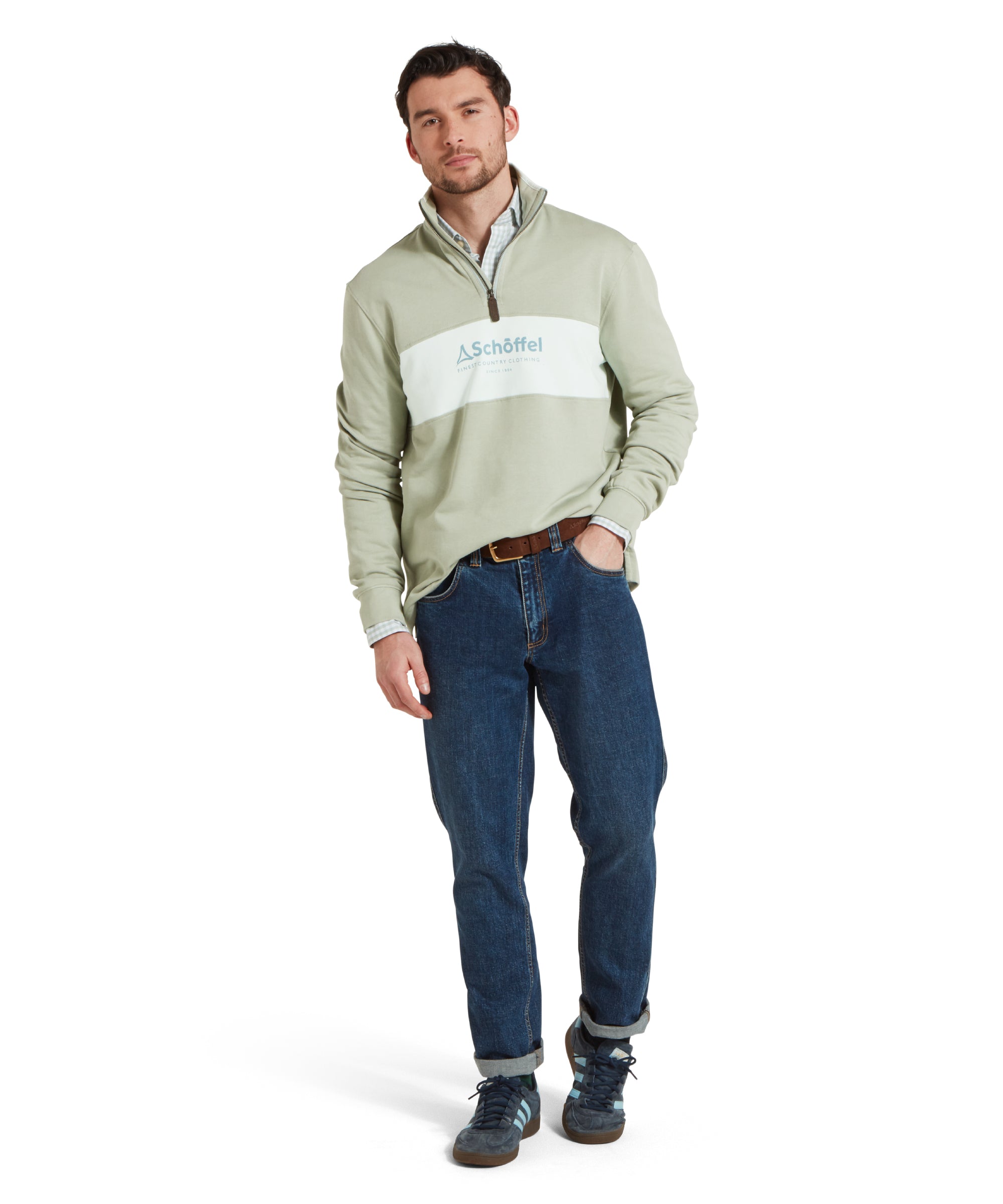 Full height image of a male model wearing a green Schöffel sweatshirt with a contrasting green stripe and large embroidered Schöffel logo across the chest. It is paired with a blue shirt, navy trainers and dark blue jeans and he is standing in a relaxed pose one hand in his pocket and he is smiling looking towards the camera.