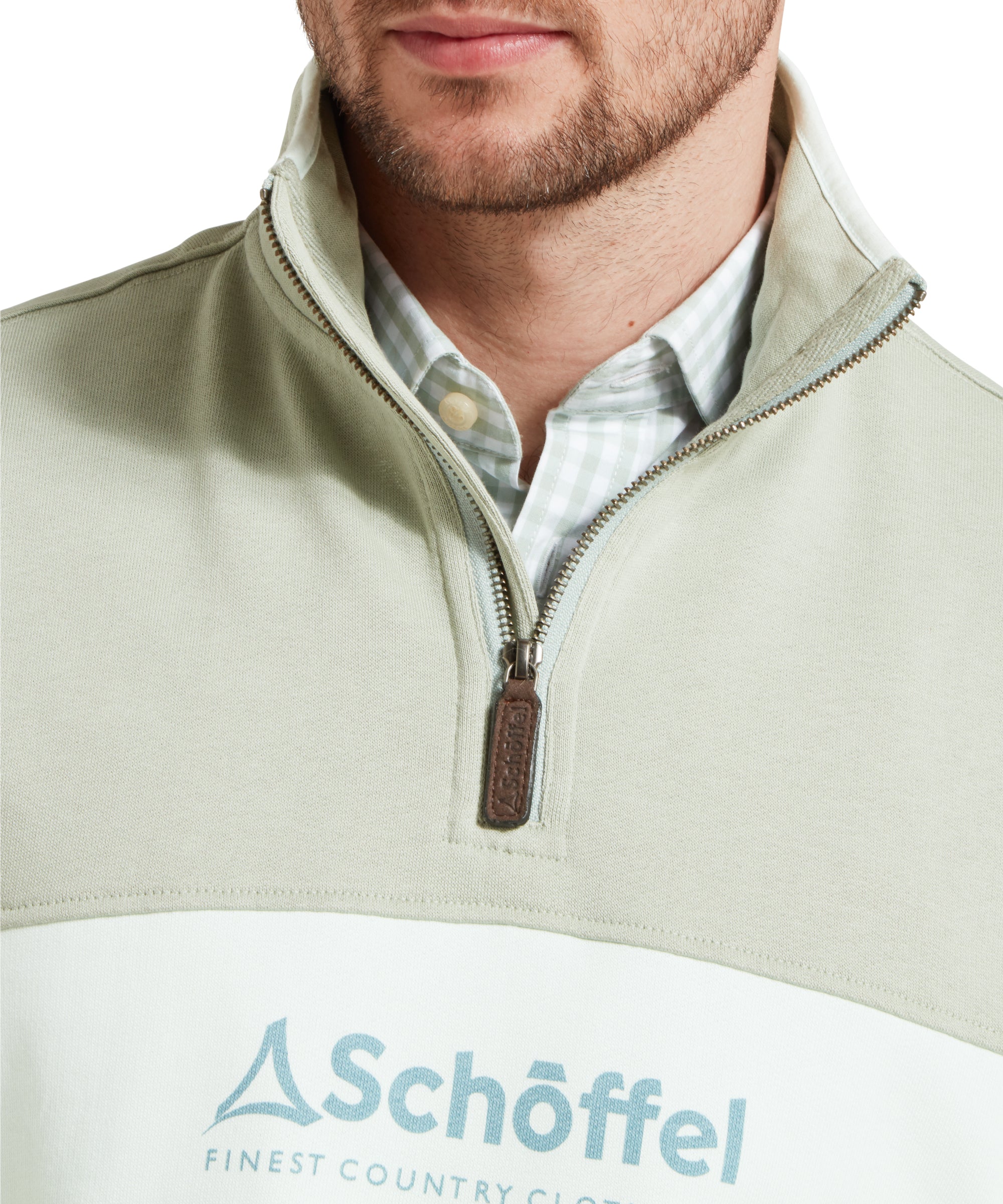 Close up image of a male model wearing a green Schöffel sweatshirt with a contrasting stripe and large embroidered Schöffel logo across the chest. It is paired with a blue gingham shirt. Image highlights quarter zip neck with pull tab.