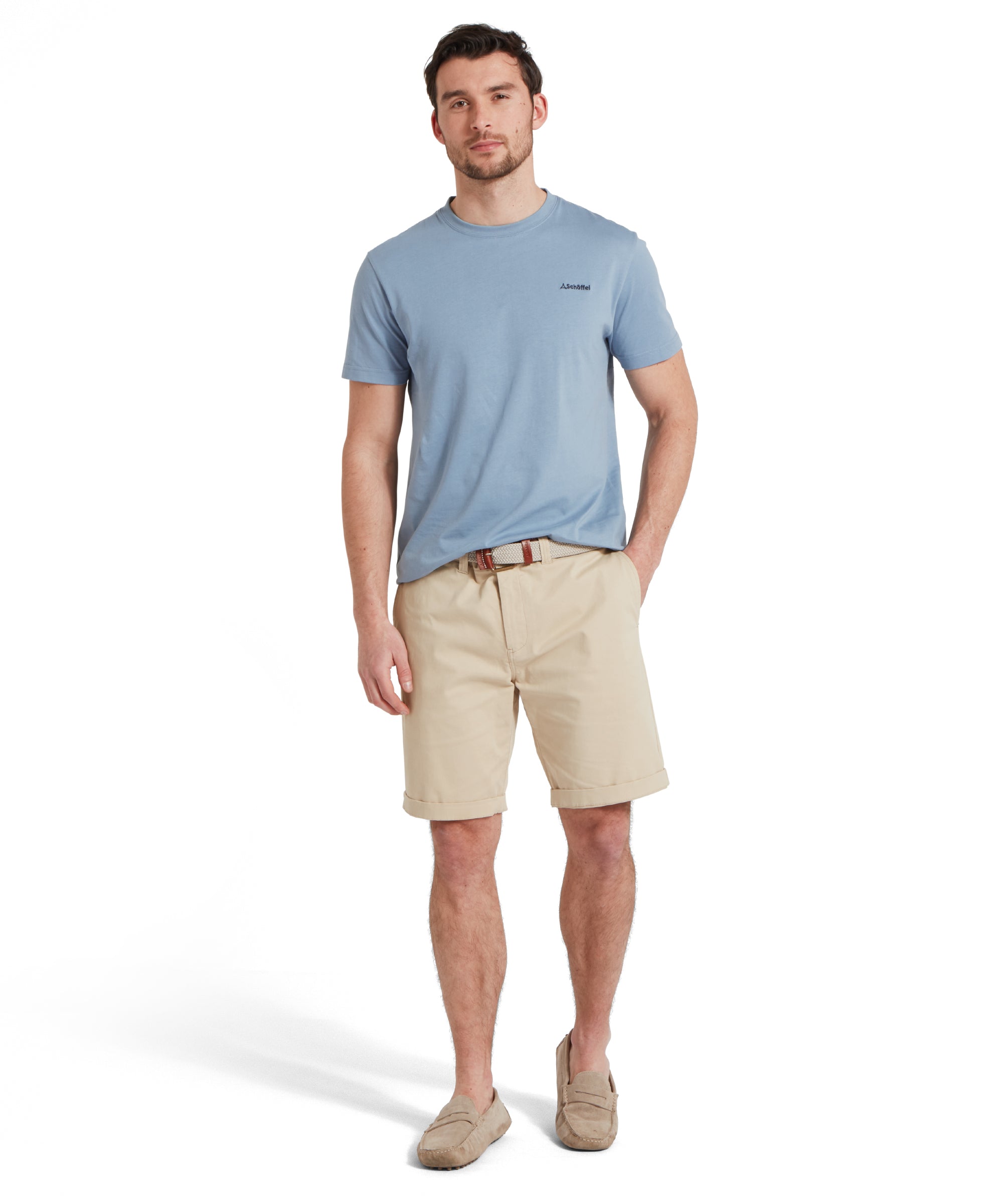 Full-height image of a male model wearing a blue Schöffel t-shirt with a small embroidered dark blue "Schöffel" logo on the chest, paired with beige shorts, tan loafers and a woven belt. He is standing in a relaxed pose with one hand in his pocket.