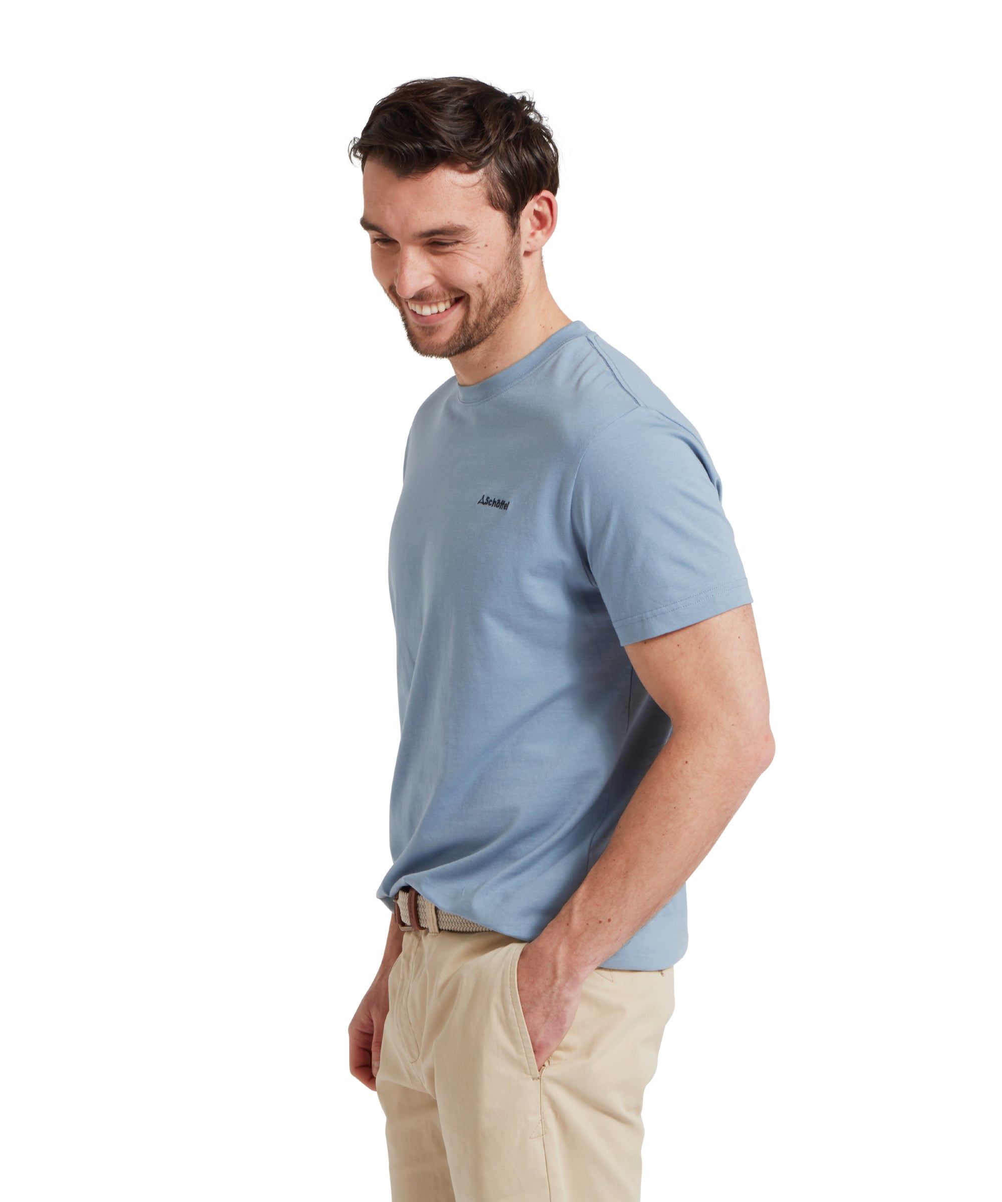 Side profile image of a male model wearing a blue Schöffel t-shirt with a small embroidered dark blue "Schöffel" logo on the chest, paired with beige shorts and a woven belt. He is standing in a relaxed pose with one hand in his pocket.