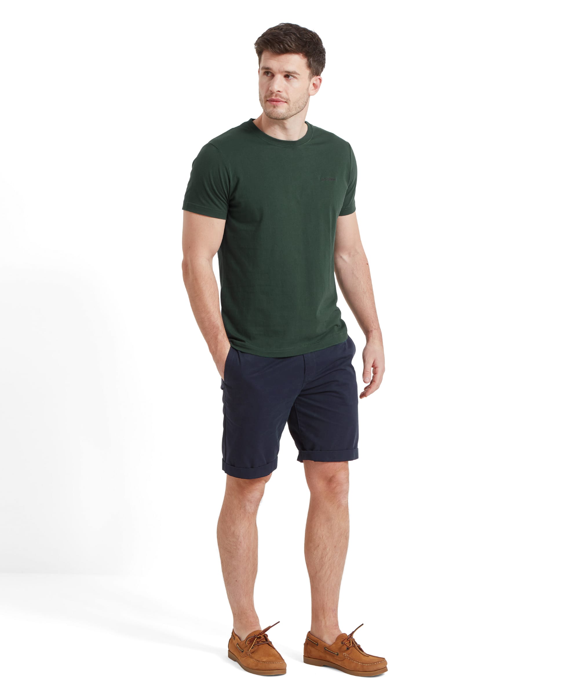 A man models a Schöffel Trevone T-Shirt for Men in Green paired with dark navy shorts and brown boat shoes. He is standing with one hand in his pocket, looking to his left.