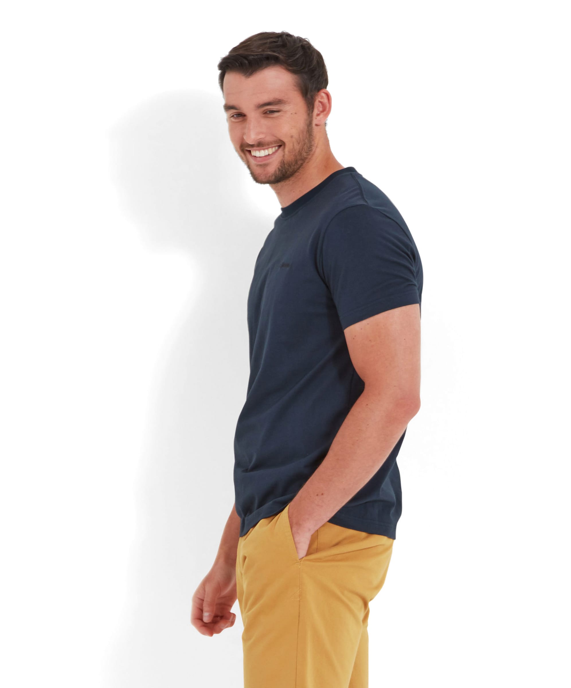 A man is smiling while wearing a Schöffel Trevone T-Shirt for Men in Navy and mustard yellow shorts, standing in a relaxed pose with hands in his pockets, facing sideways.