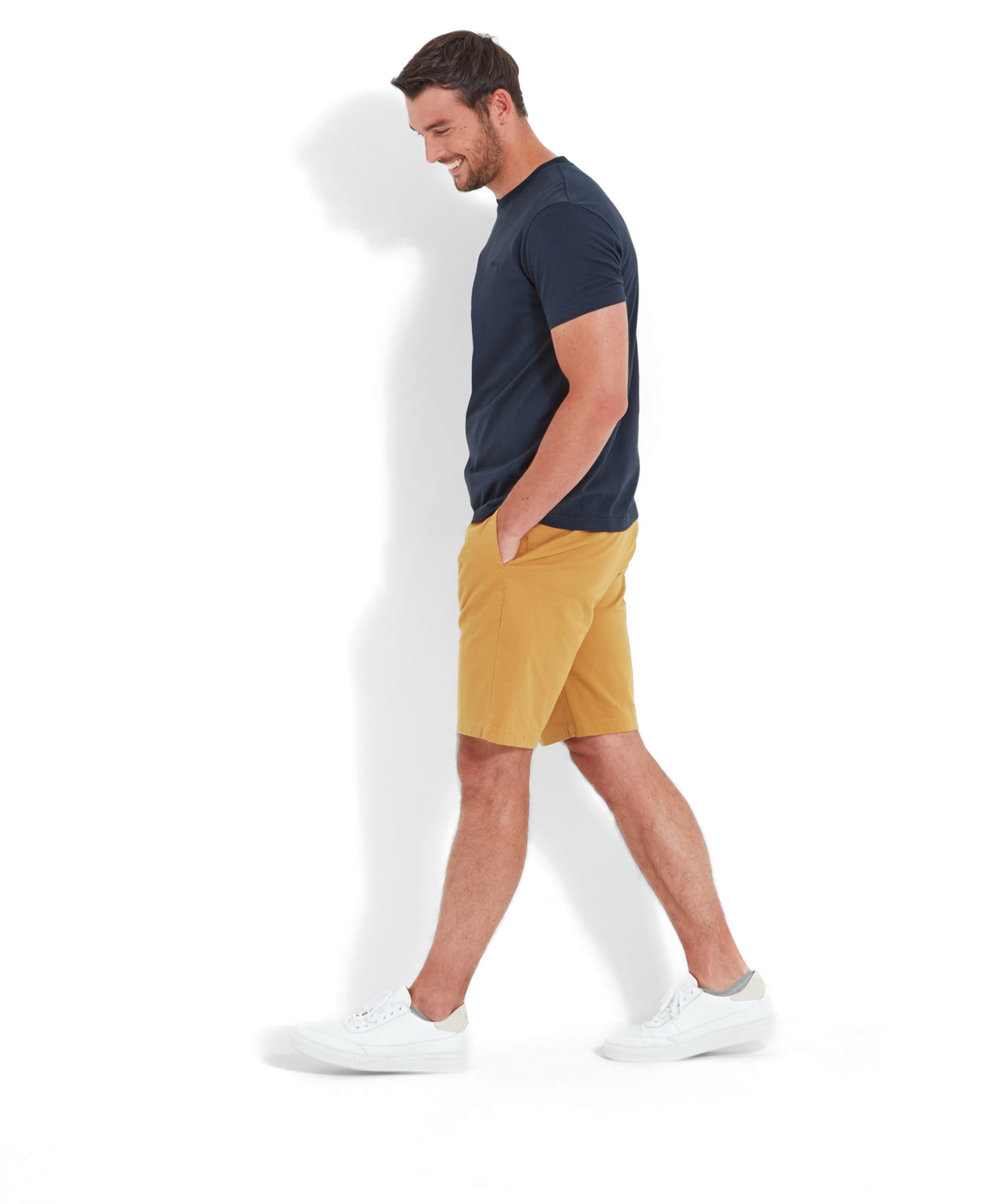 The same man walks in the Schöffel Trevone T-Shirt for Men in Navy and mustard yellow shorts, looking down with a slight smile, captured in a side view.