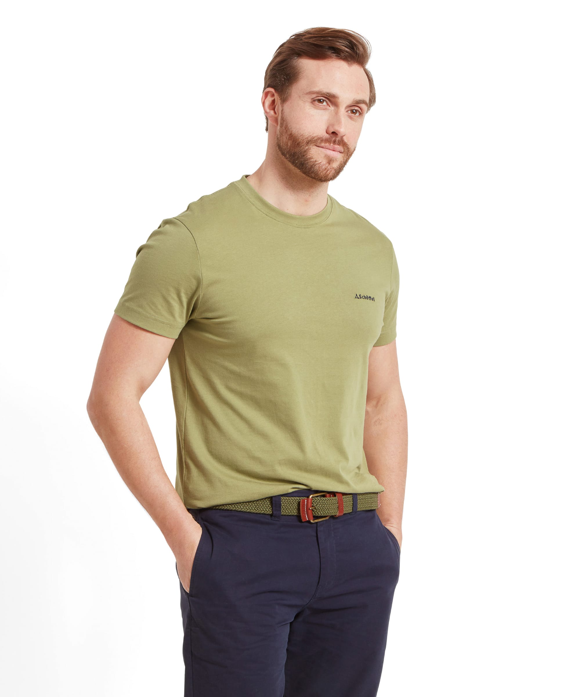 A man is standing against a white background, wearing a Schöffel Trevone T-Shirt for Men in Olive Green and dark navy shorts. The T-shirt is plain with a subtle logo on the left chest. The man has a neatly groomed beard and styled brown hair. He looks slightly off to the side with a neutral expression.