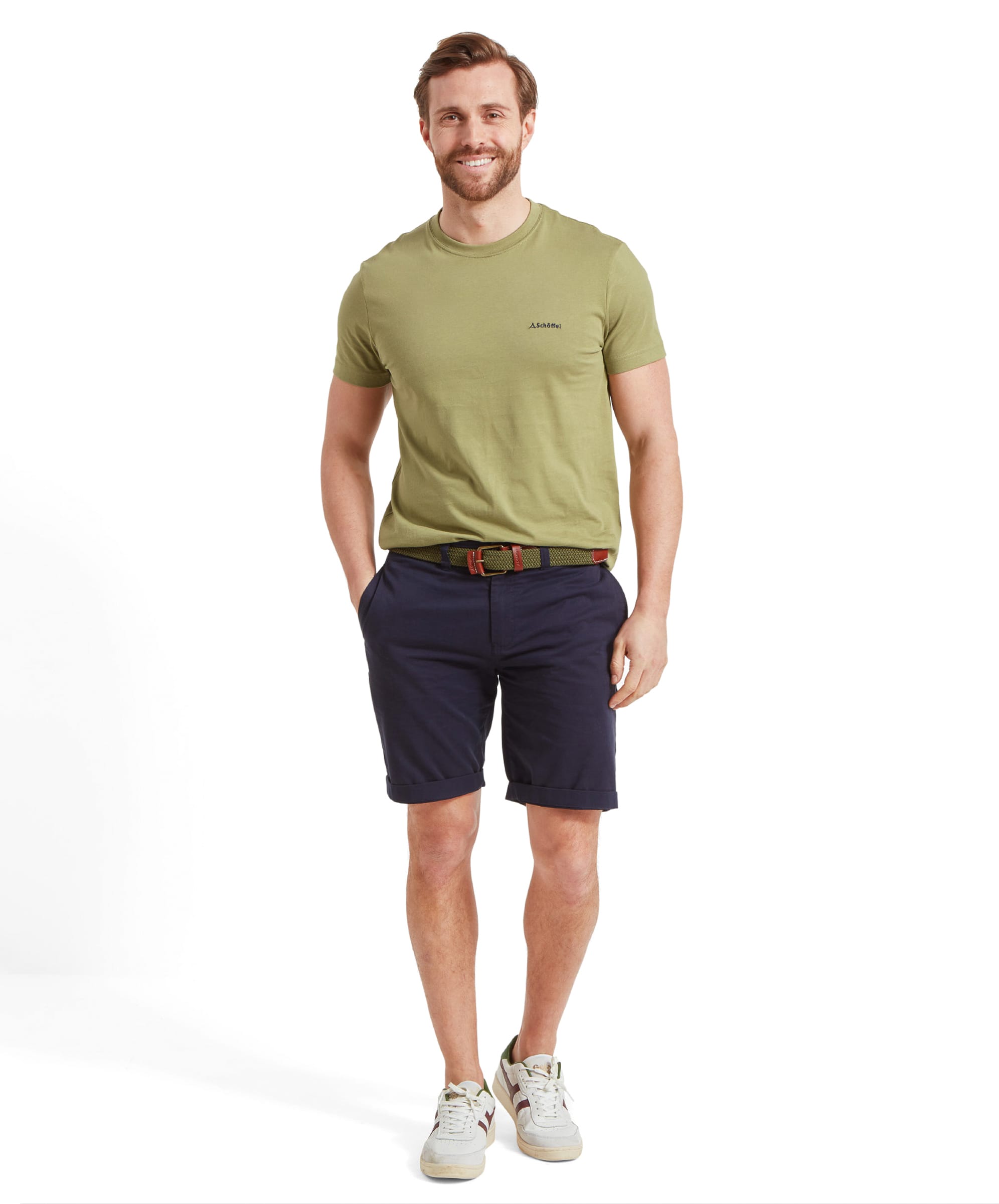 The man is posing full-length in the same outfit, smiling slightly. The Schöffel Trevone T-Shirt for Men in Olive Green is tucked into his navy shorts, and he is wearing white trainers. His hands are in his pockets, giving a casual look.