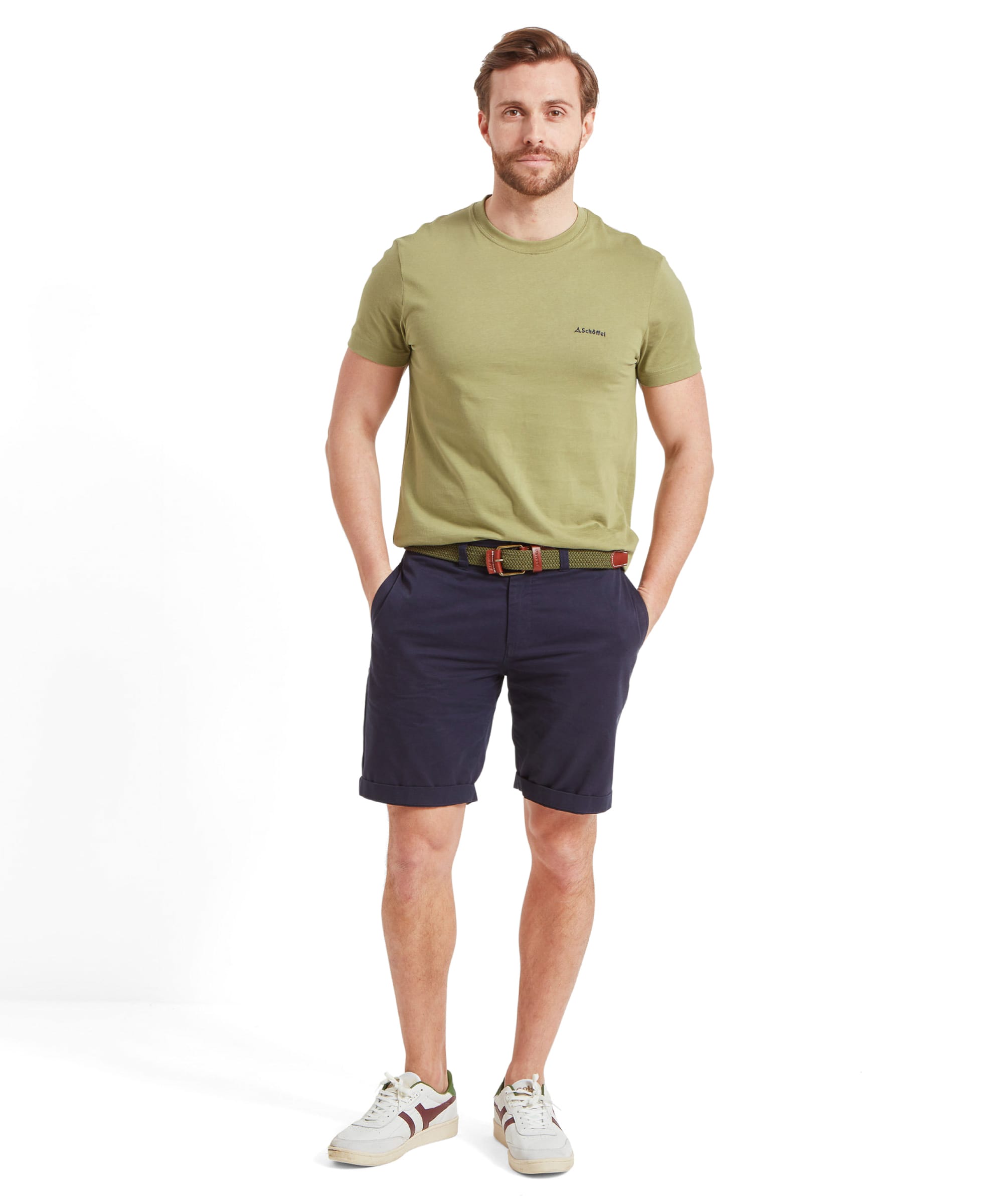 The man poses with his hands in his pockets, looking down slightly, showcasing a relaxed and casual stance. The outfit is the same as before, with a Schöffel Trevone T-Shirt for Men in Olive Green and navy shorts.