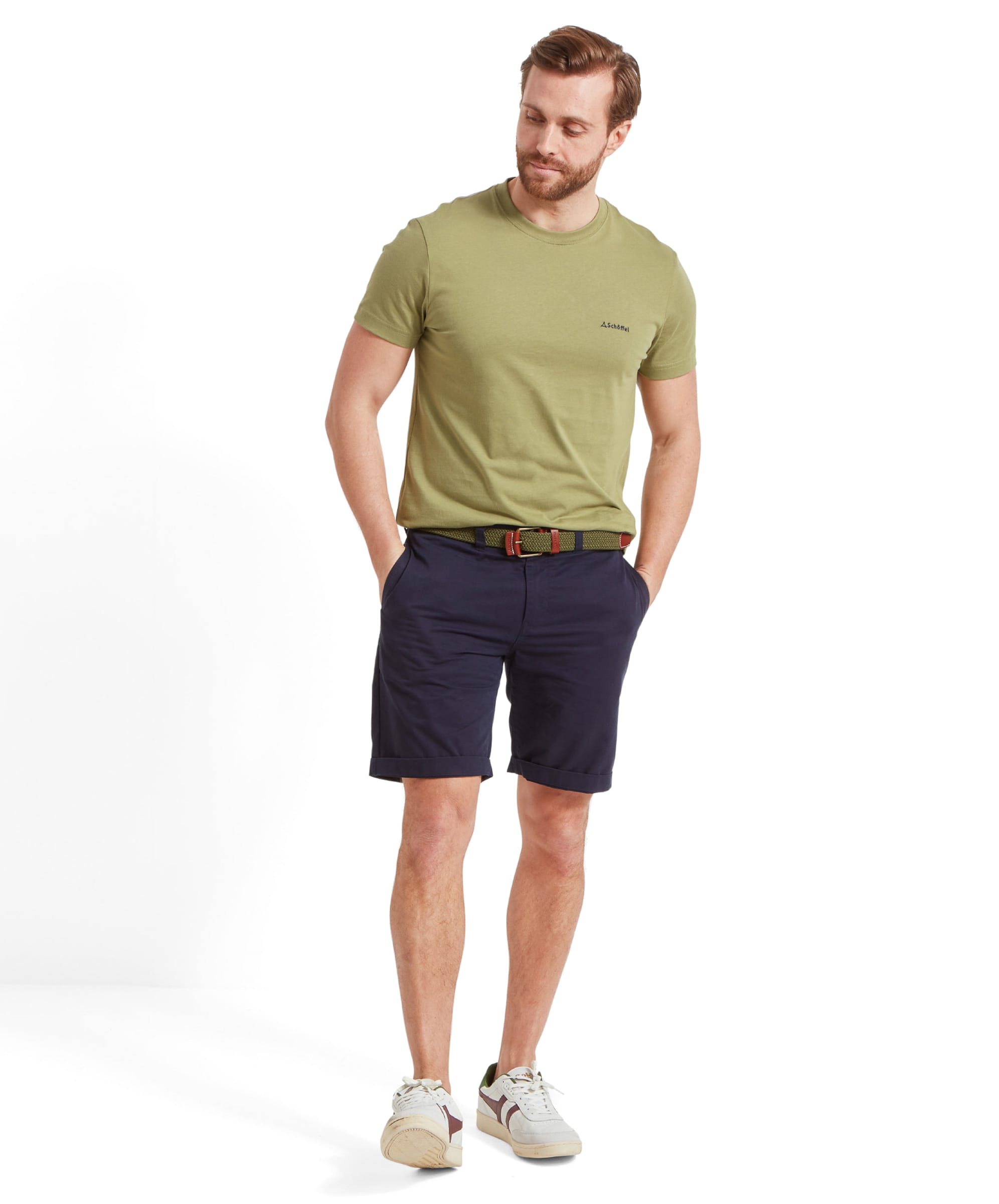 A man is standing wearing a Schöffel Trevone T-Shirt for Men in Olive Green, paired with navy shorts. He is also wearing a matching belt and white rtainers. The shirt features a small Schöffel logo on the chest.