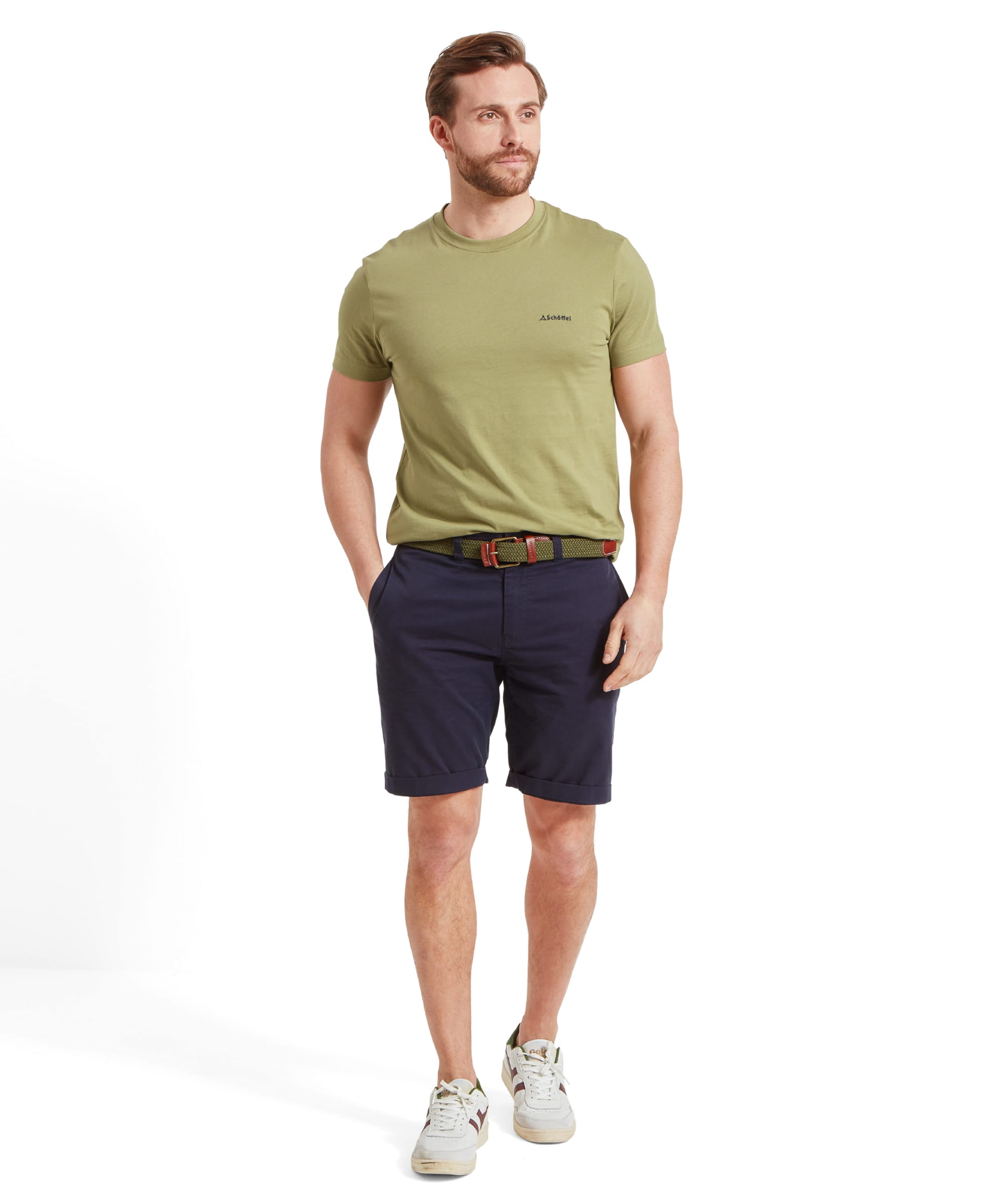 The same man is standing in a relaxed pose, looking down with his hands in his pockets. He is dressed in a Schöffel Trevone T-Shirt for Men in Olive Green, navy shorts, and white trainers. The Schöffel logo is visible on the chest.