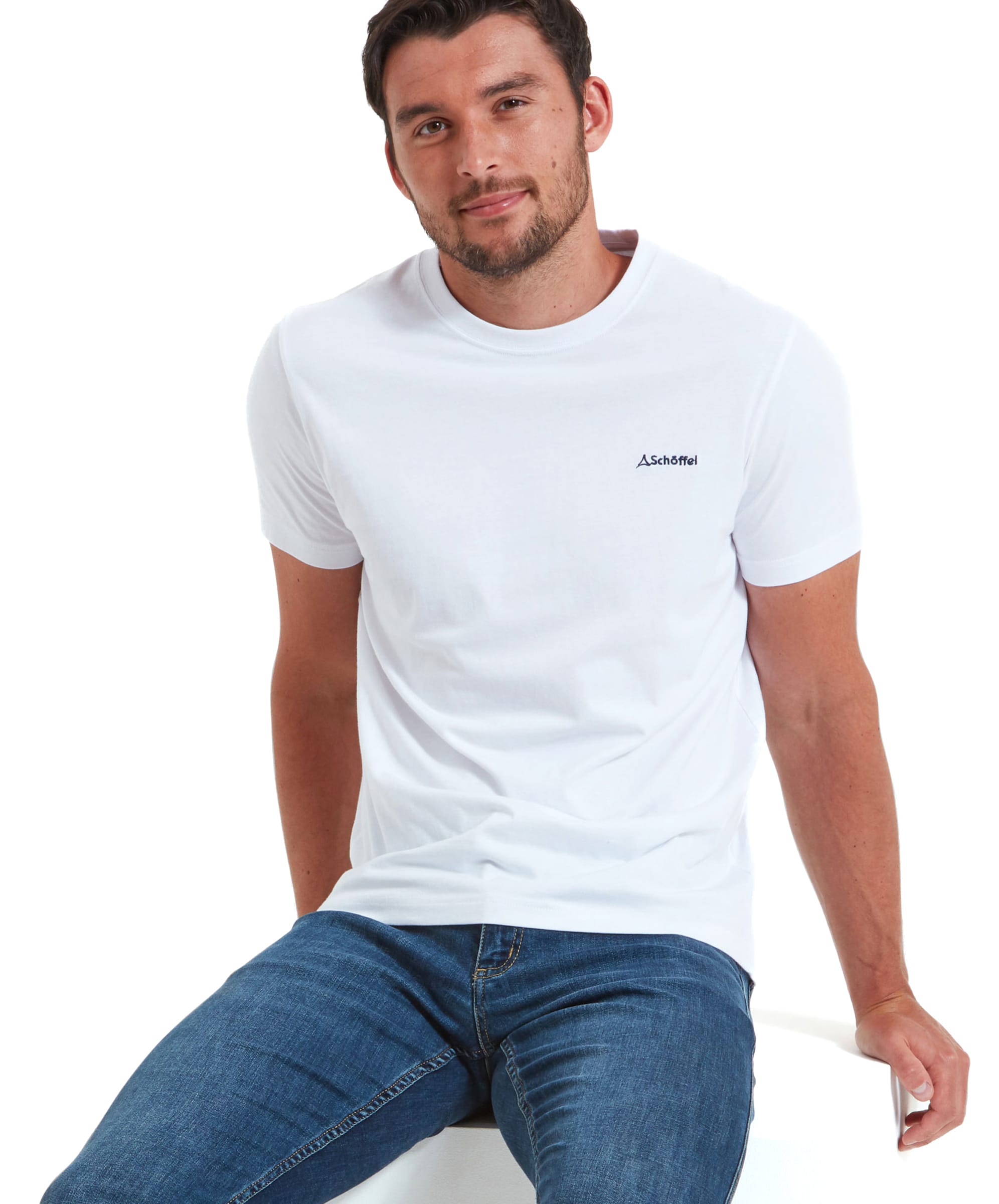 A male model sitting casually in a Schöffel Trevone T-Shirt for Men in White and blue jeans, smiling at the camera. The t-shirt has a small embroidered logo on the chest.