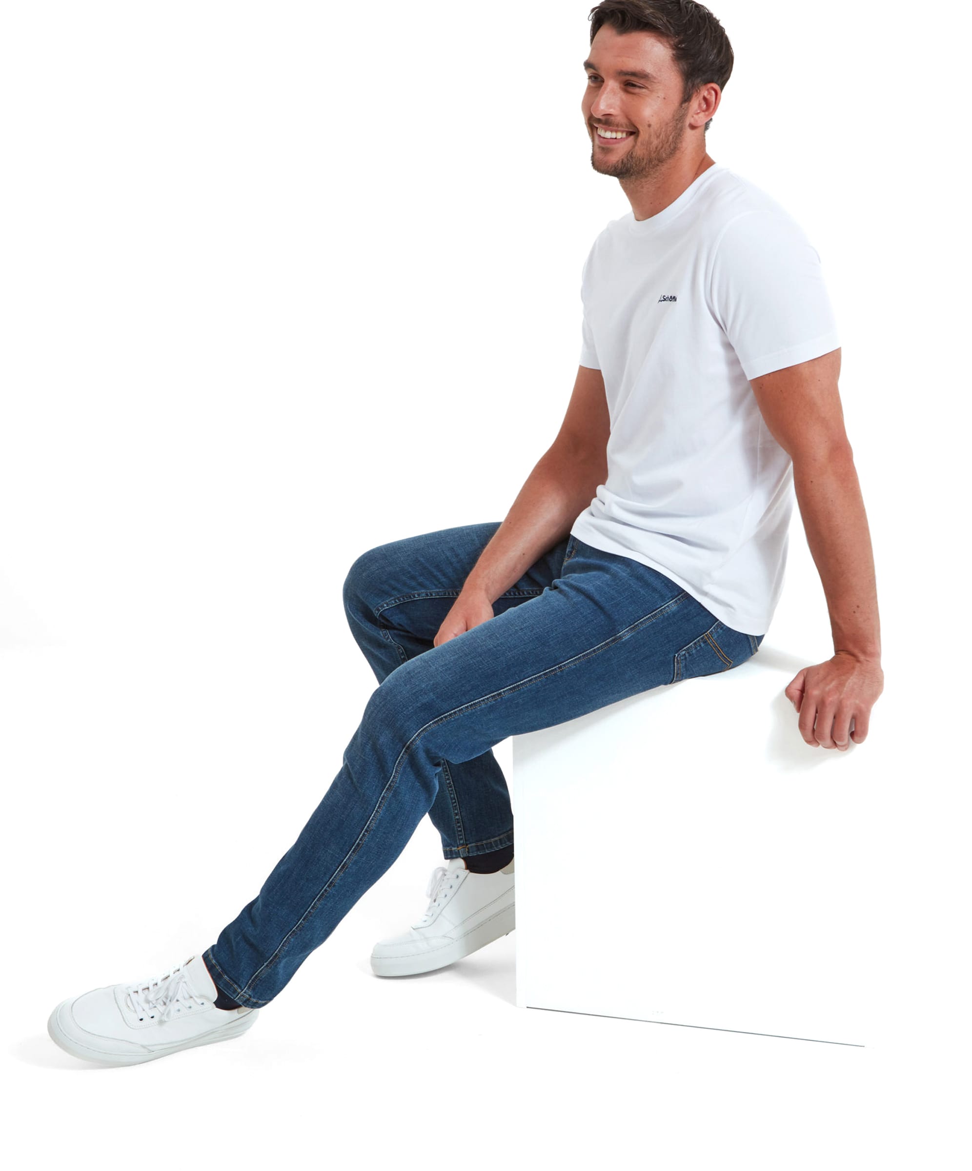 The model is sitting on a white block, showcasing the full outfit with Schöffel Trevone T-Shirt for Men in White, blue jeans, and white trainers.