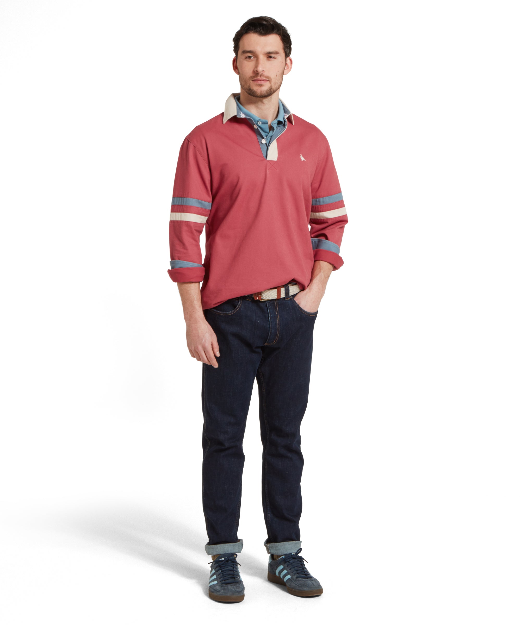 A full height image of a male model wearing a red Schöffel deck shirt with a small embroidered bird logo on the chest and blue and beige stripes on the arms. It is paired with dark denim jeans, blue polo shirt, beige belt and navy trainers. He is standing in a relaxed pose with one hand in his pocket.