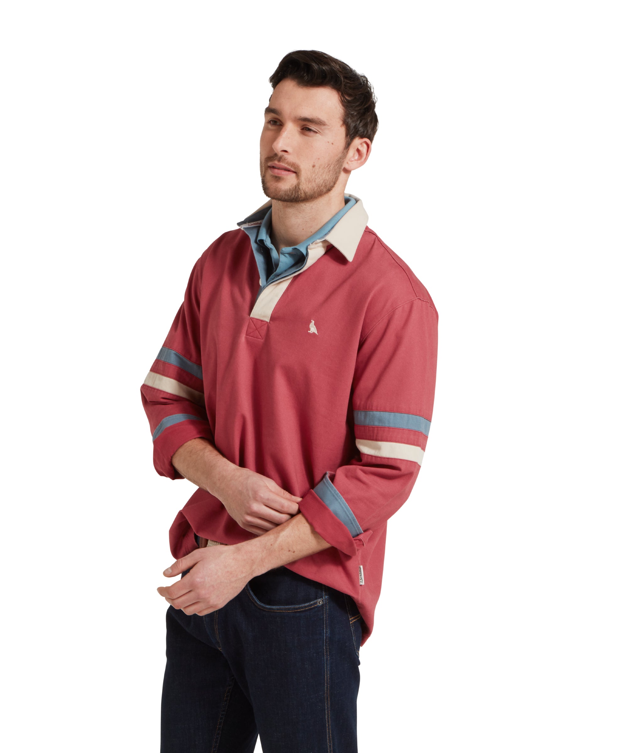 A side image of a male model wearing a red Schöffel deck shirt with a small embroidered bird logo on the chest and blue and beige stripes on the arms. It is paired with dark denim jeans, blue polo shirt and beige belt.