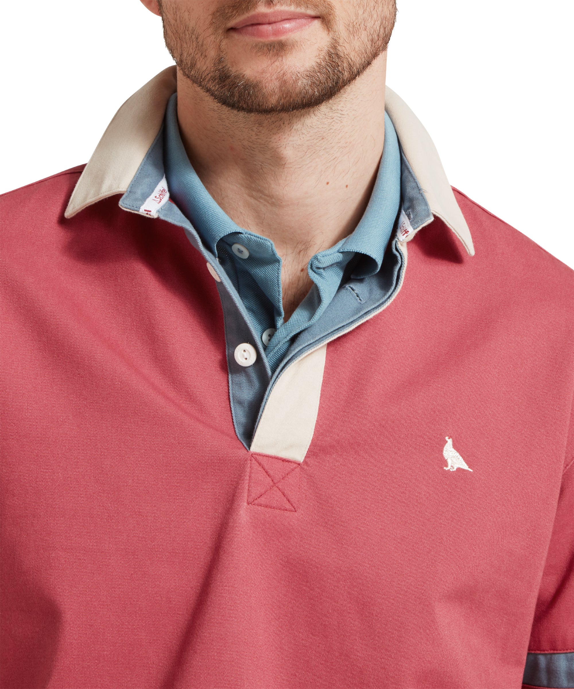 A close up image of a male model wearing a red Schöffel deck shirt with a small embroidered bird logo on the chest and blue and beige stripes on the arms. It is paired a blue polo shirt. The image highlights the collar of the shirt with light blue inner material and 2 buttons.