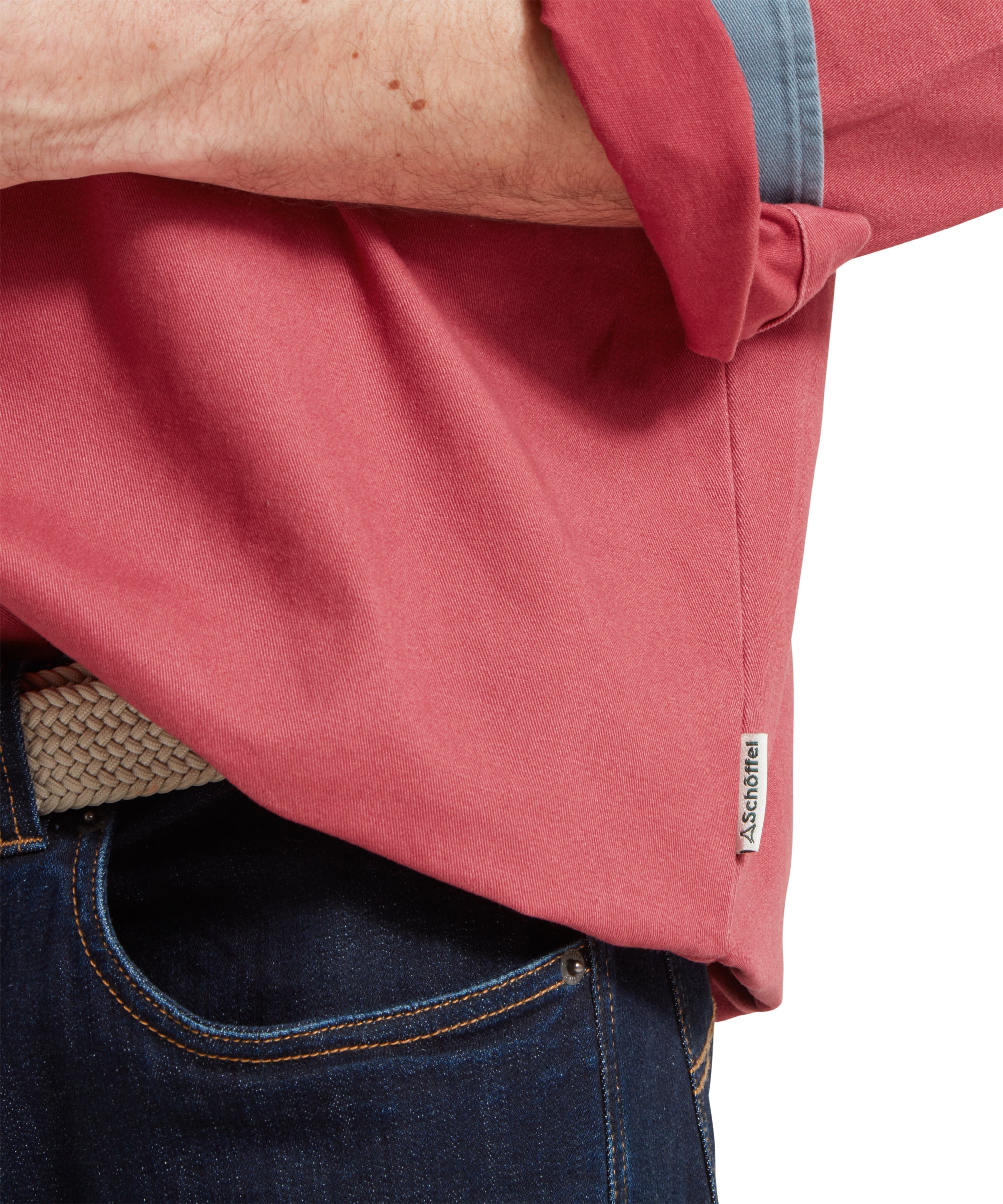 A close up image of a male model wearing a red Schöffel deck shirt with a small embroidered bird logo on the chest and blue and beige stripes on the arms. It is paired a blue polo shirt. The image highlights the hem of the shirt with small white 'Schöffel' logo tag.