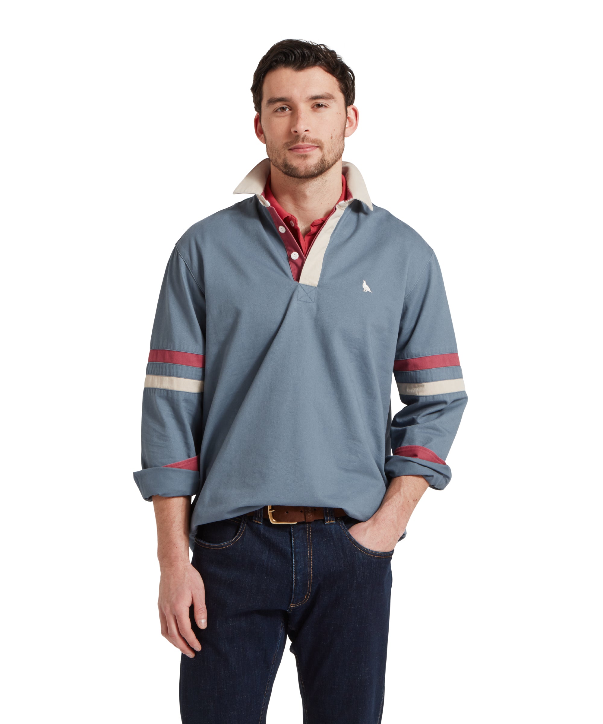 Half height image of a male model wearing a blue Schöffel sweatshirt with red and white stripe detailing on the sleeves and small embroidered bird logo on the chest. It is paired with a red polo and dark jeans. He is standing in a relaxed pose with one hand in his pocket.