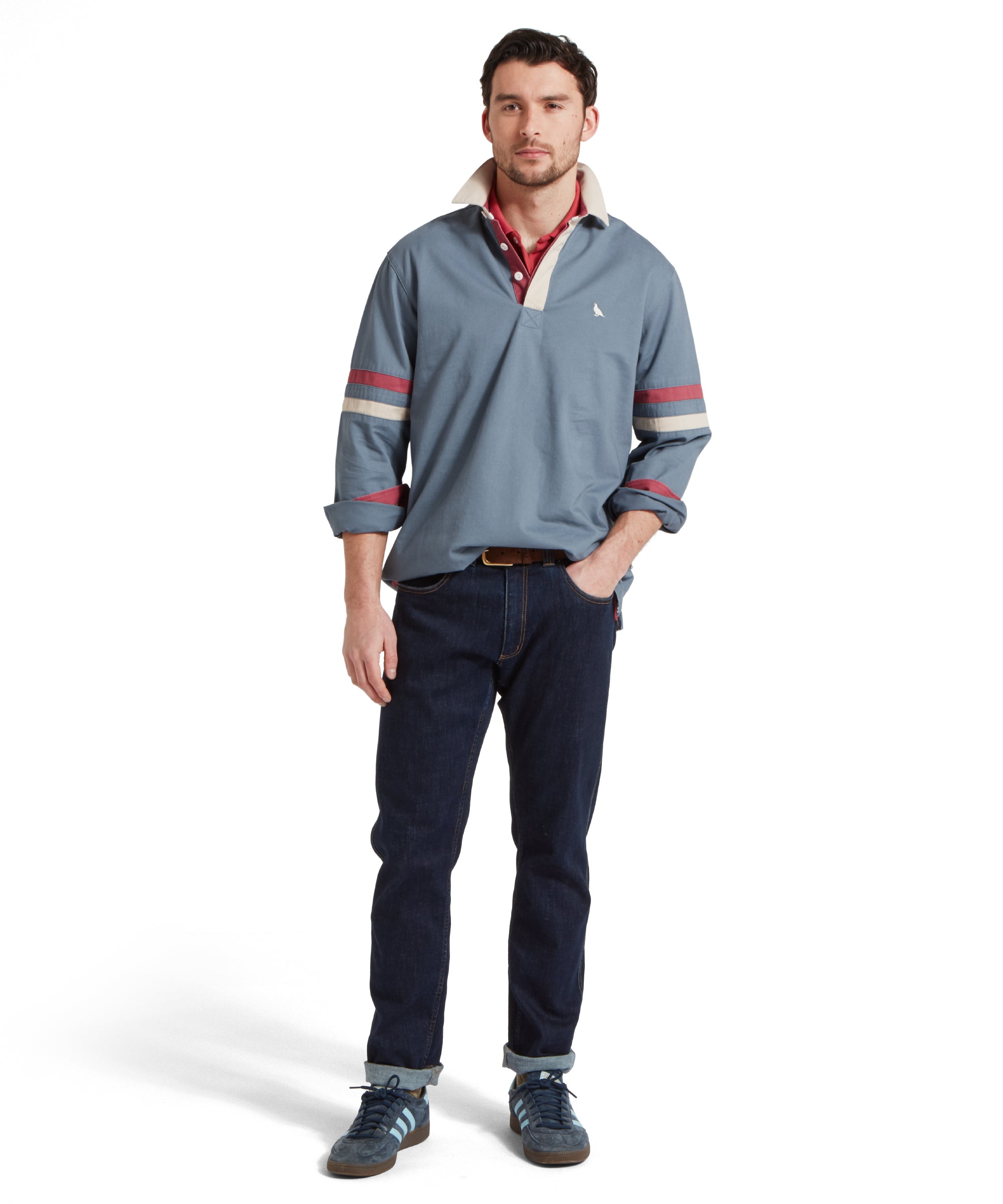 Full height image of a male model wearing a blue Schöffel sweatshirt with red and white stripe detailing on the sleeves and small embroidered bird logo on the chest. It is paired with a red polo, dark jeans and blue trainers. He is standing in a relaxed pose with one hand in his pocket.