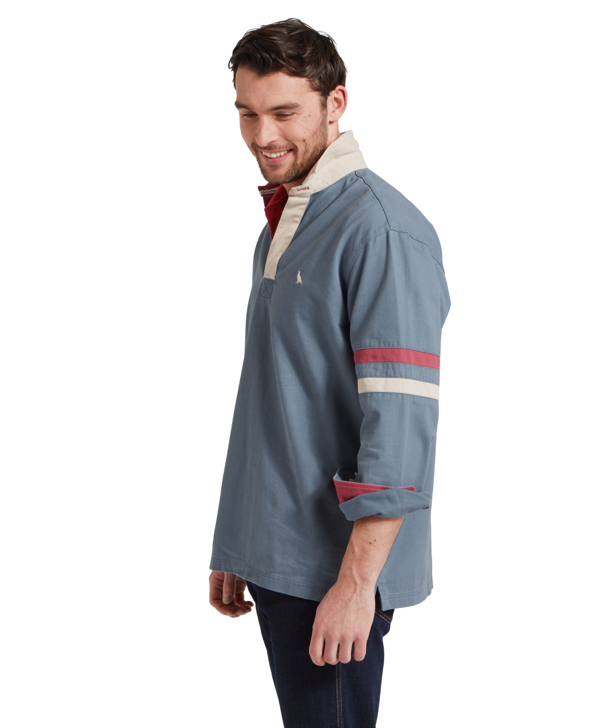 Side view of a male model wearing a blue Schöffel sweatshirt with red and white stripe detailing on the sleeves and small embroidered bird logo on the chest. It is paired with a red polo and dark jeans.