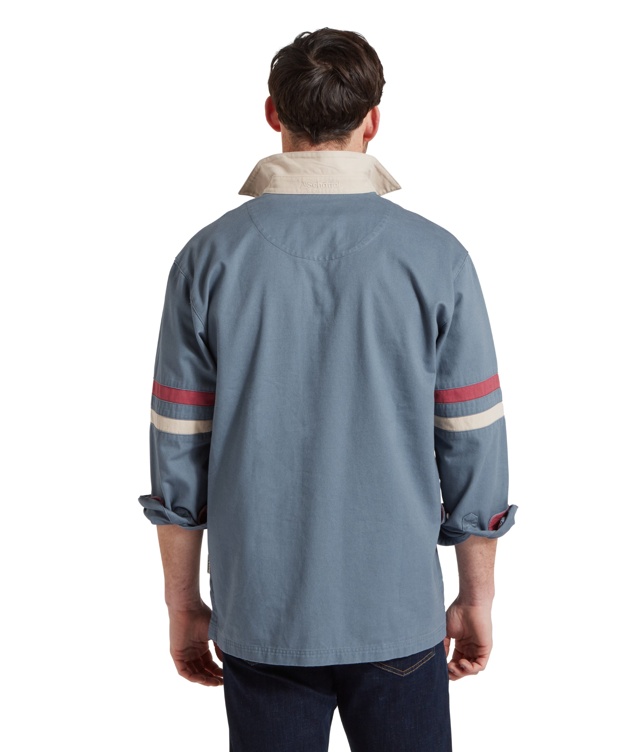 Back image of a male model wearing a blue Schöffel sweatshirt with red and white stripe detailing on the sleeves and small embroidered bird logo on the chest. It is paired with a red polo and dark jeans. He is standing in a relaxed pose with his hands by his side.