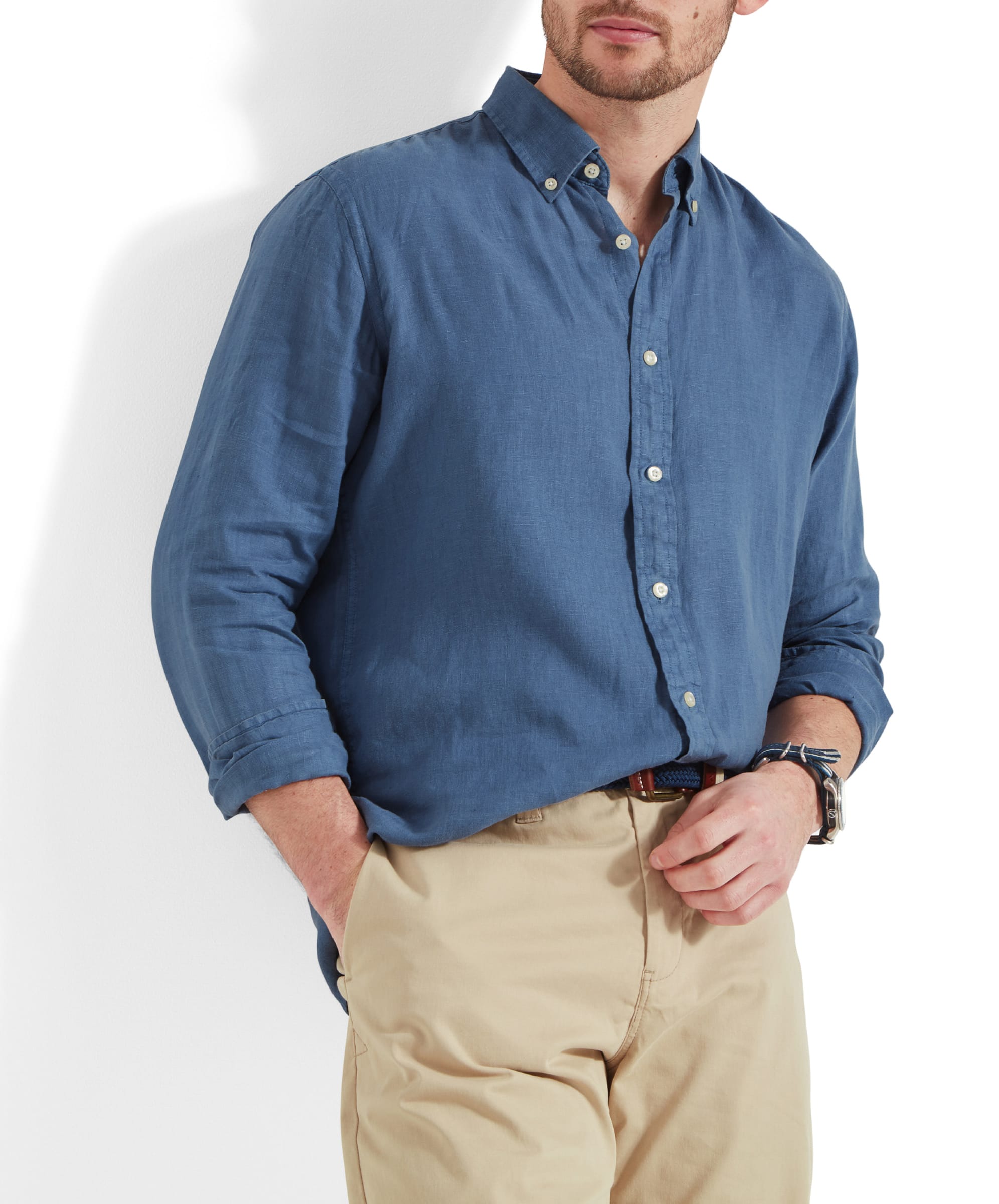 A man dressed in a Schöffel Walsingham Linen Shirt for Men in Blue with long sleeves, buttoned up, and tucked into beige shorts. He has his hands in his pockets, displaying a relaxed yet stylish pose. The shirt's soft, breathable fabric and well-crafted details, like the button-down collar, are highlighted.