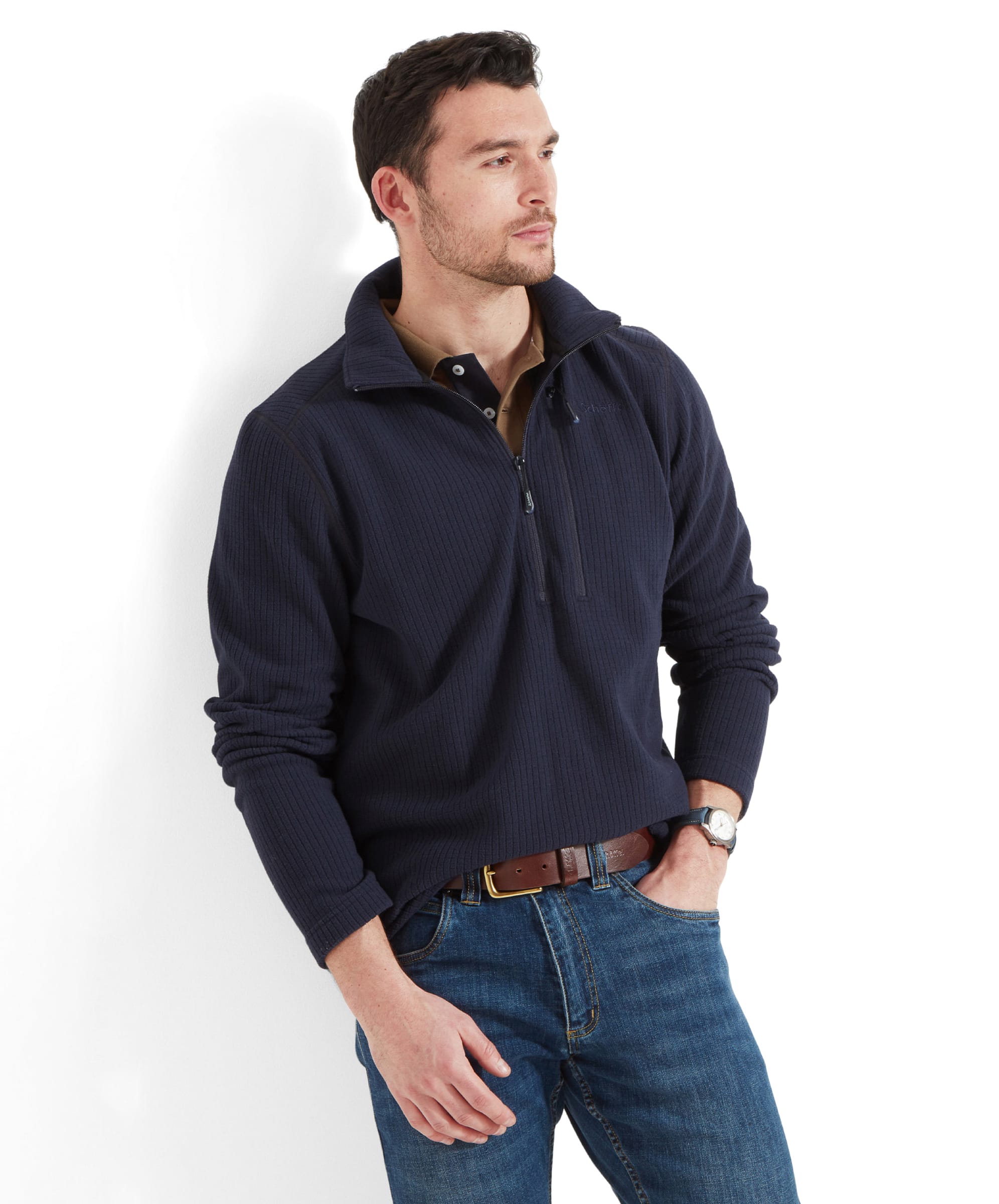 A male model is wearing a Schöffel Welland Lightweight Fleece for Men in Blue with a half-zip design. The fleece has a textured, ribbed pattern and is paired with jeans. The model is accessorised with a brown leather belt and a wristwatch. The model's look is casual and relaxed, standing against a white background.