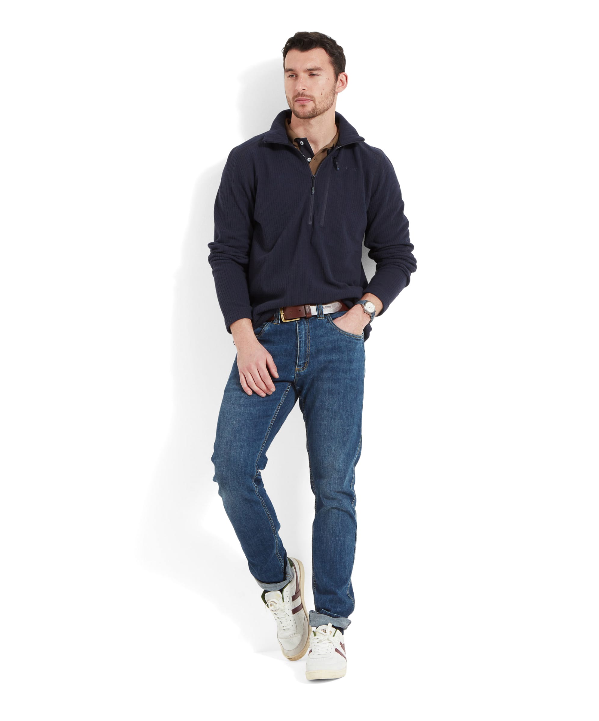 The same male model is standing casually, wearing the same Schöffel Welland Lightweight Fleece for Men in Blue with a half-zip design. The fleece features a textured pattern and is paired with blue jeans. The model accessorizes with a brown leather belt, wristwatch, and white trainers, exuding a comfortable and stylish vibe.