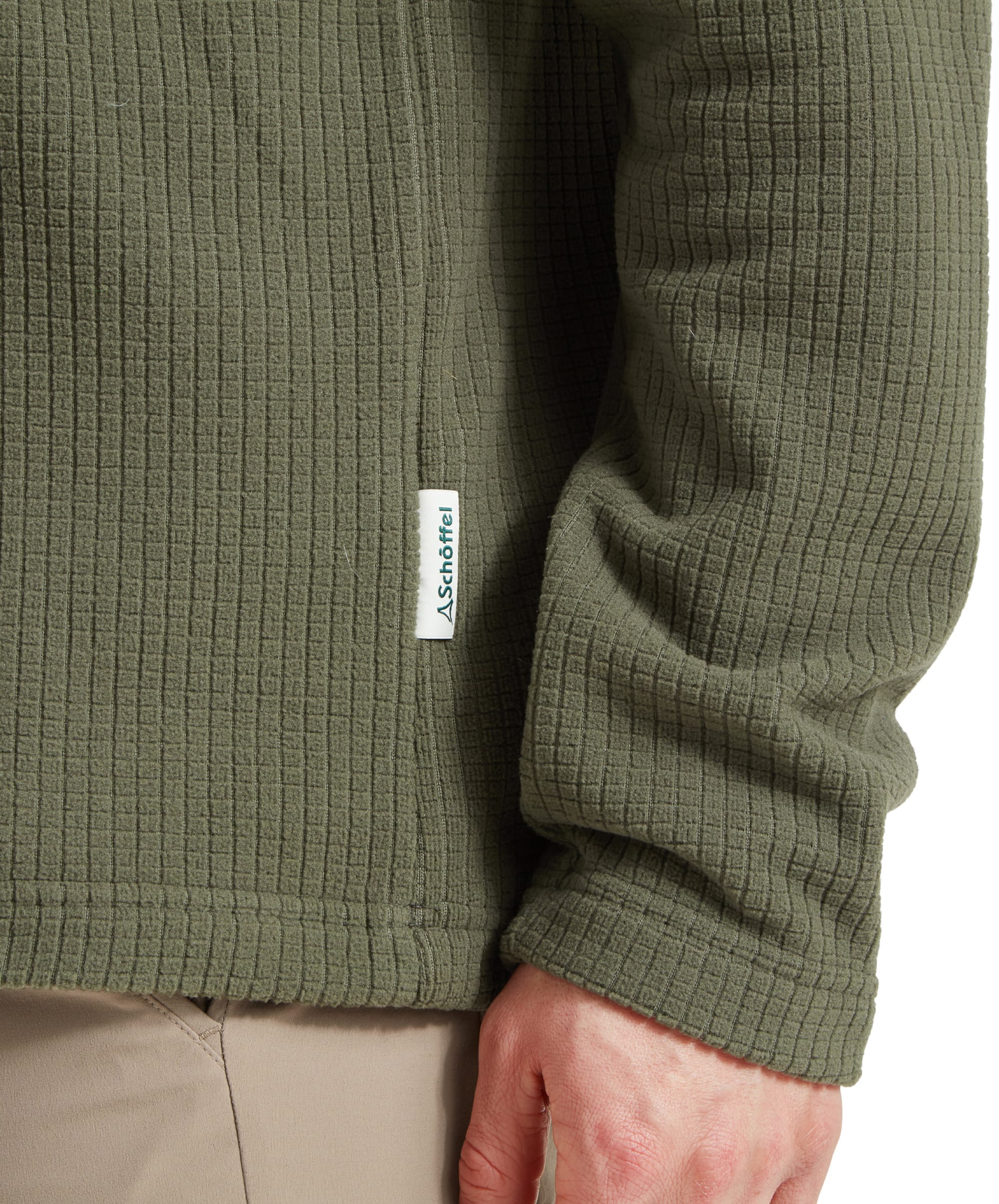 A close-up shot focusing on the side pip detail of the Schöffel Welland Lightweight Fleece for Men in Green, showcasing the Schöffel brand tag and the texture of the fabric. The image emphasizes the attention to detail in the garment's construction.