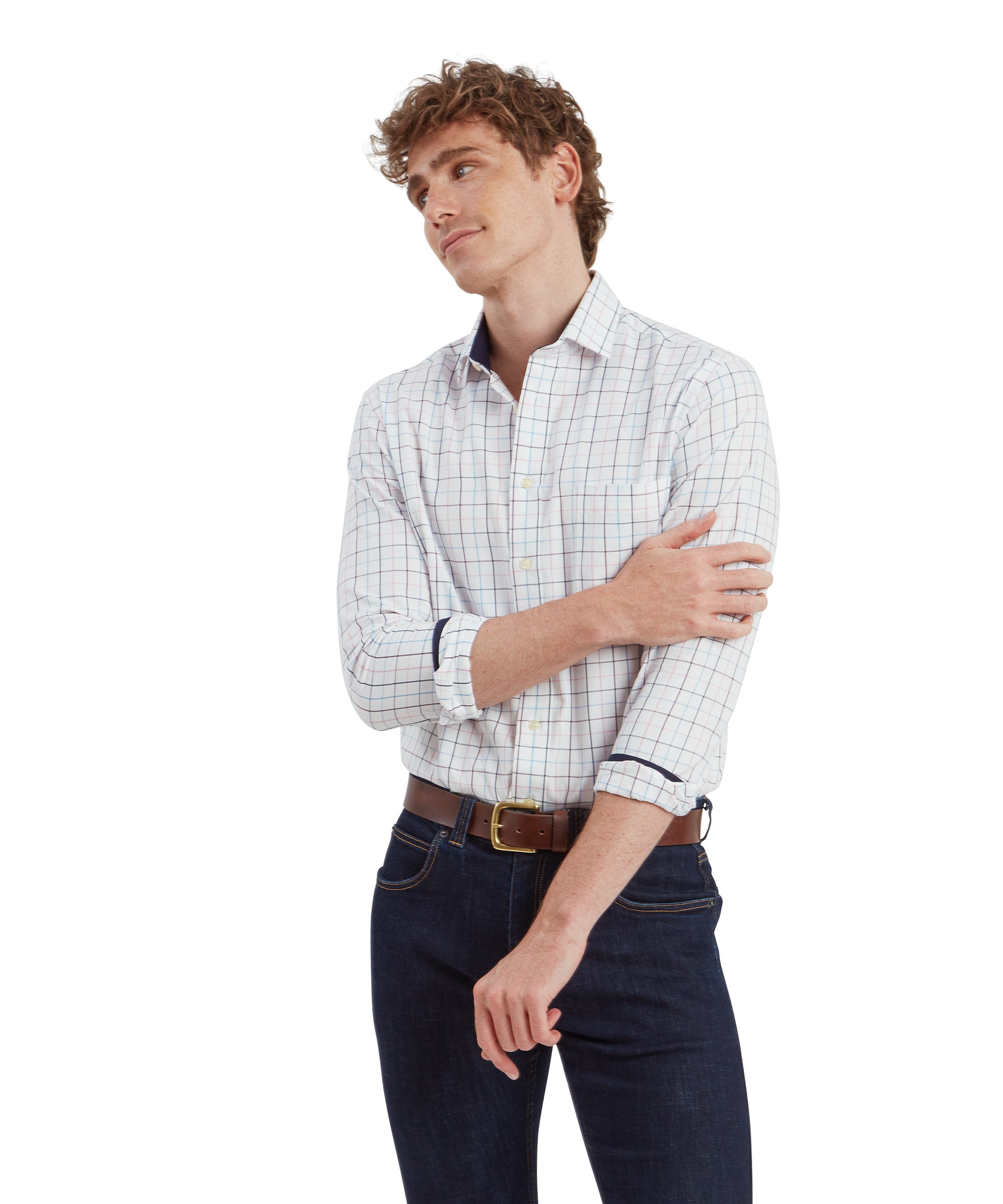 A young man with curly light brown hair is wearing a Schöffel Wells Tailored Shirt for Men in Red with a grid pattern of thin red, blue, and light brown lines. The shirt is neatly tucked into dark blue jeans, accessorised with a brown leather belt. He is standing in a relaxed pose, with one arm across his body, lightly touching the opposite arm, and has a gentle smile on his face.