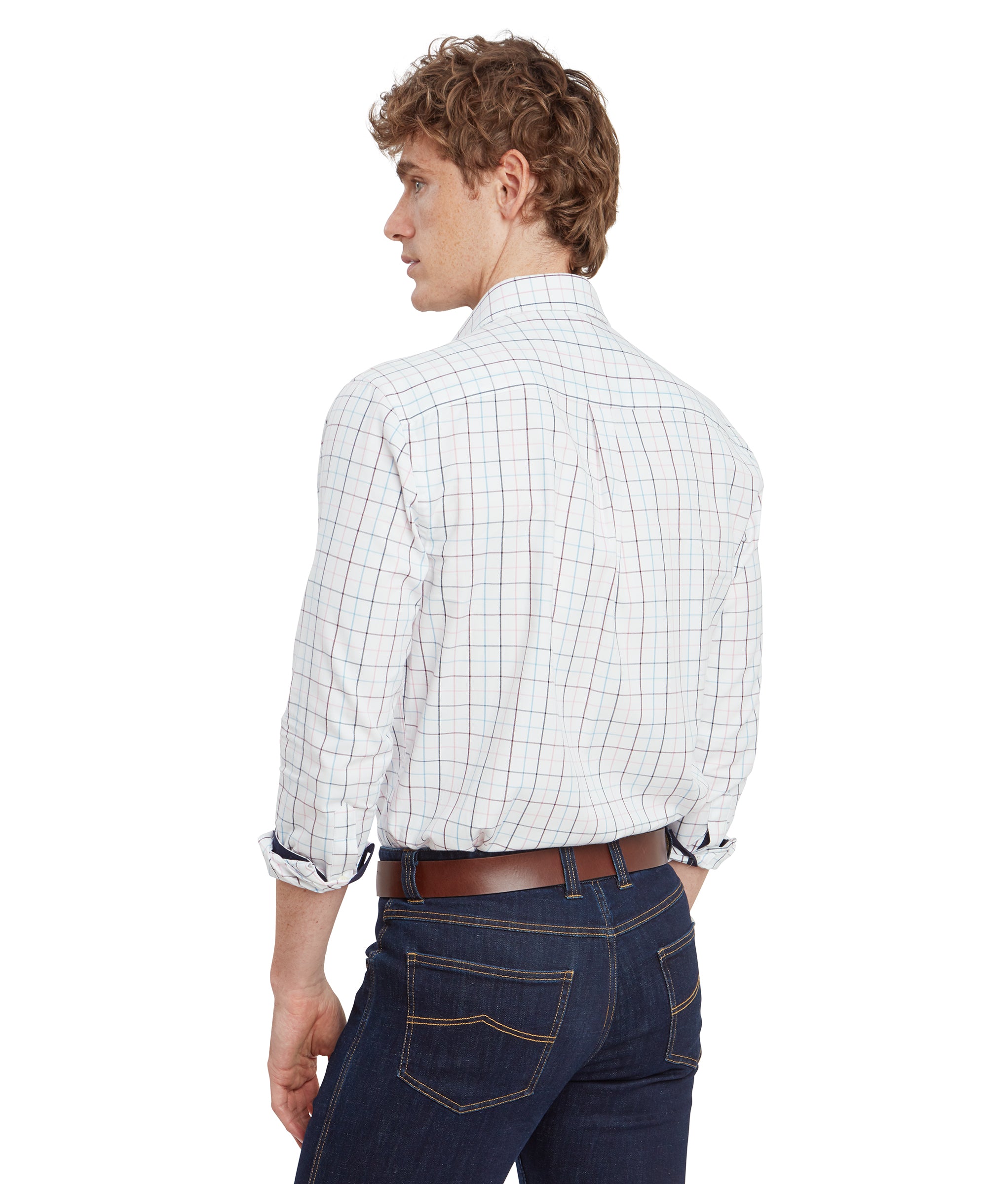 A rear view of the young man in the Schöffel Wells Tailored Shirt for Men in Red. The back of the shirt features a neat yoke and pleats, providing a tailored fit. The dark blue jeans are well-fitted, and his brown belt complements the outfit. The sleeves of the shirt are rolled up just above the elbows, revealing the navy blue inner lining.