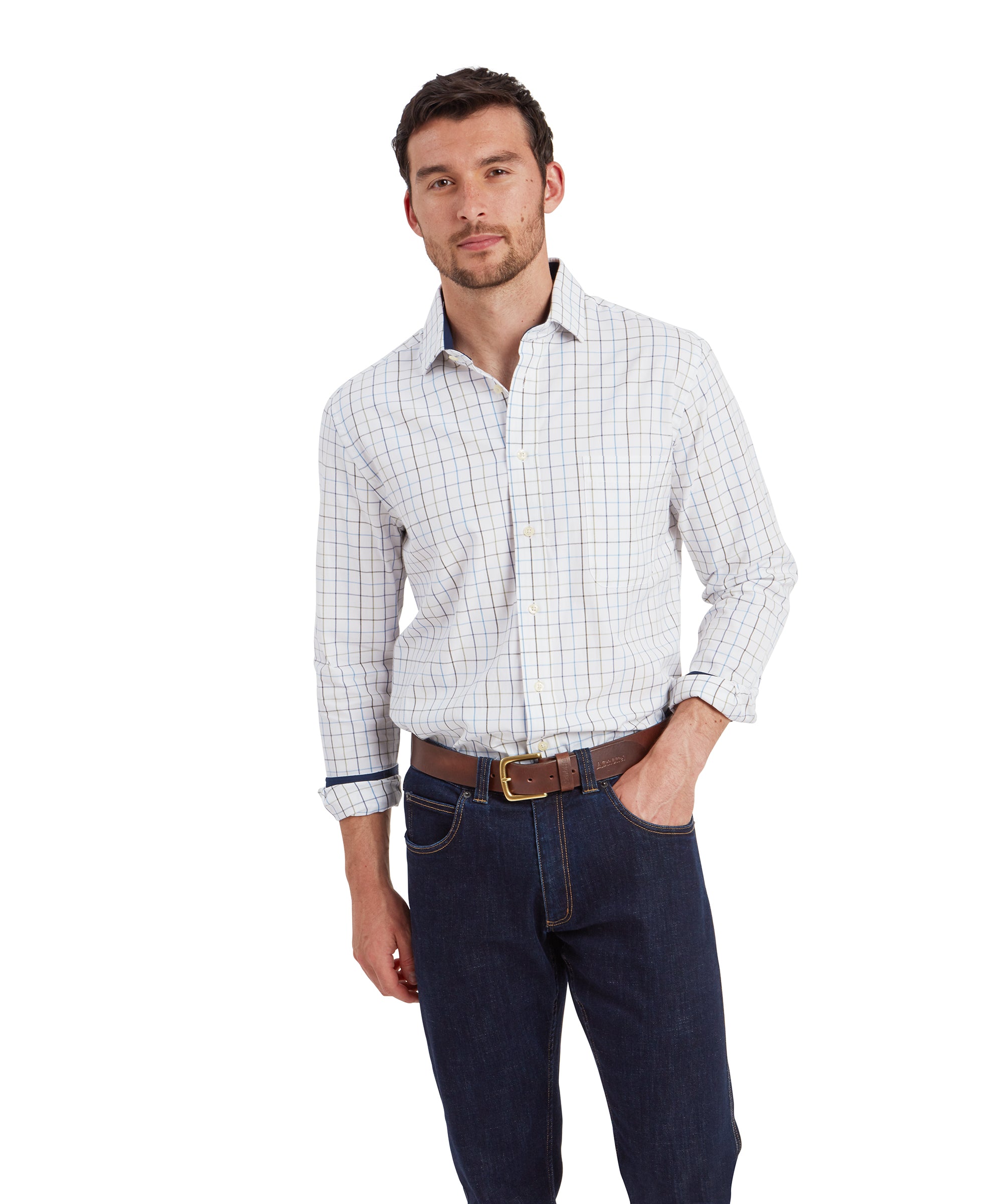 The man is standing with a neutral expression, wearing a Schöffel Wells Tailored Shirt for Men in Blue with a checkered pattern in light tones of blue, brown, and white. The shirt has a button-down collar and a chest pocket on the left side. He is also wearing dark blue jeans and a brown leather belt. The sleeves are rolled up, and the man is casually resting one hand in his pocket while the other is slightly bent.