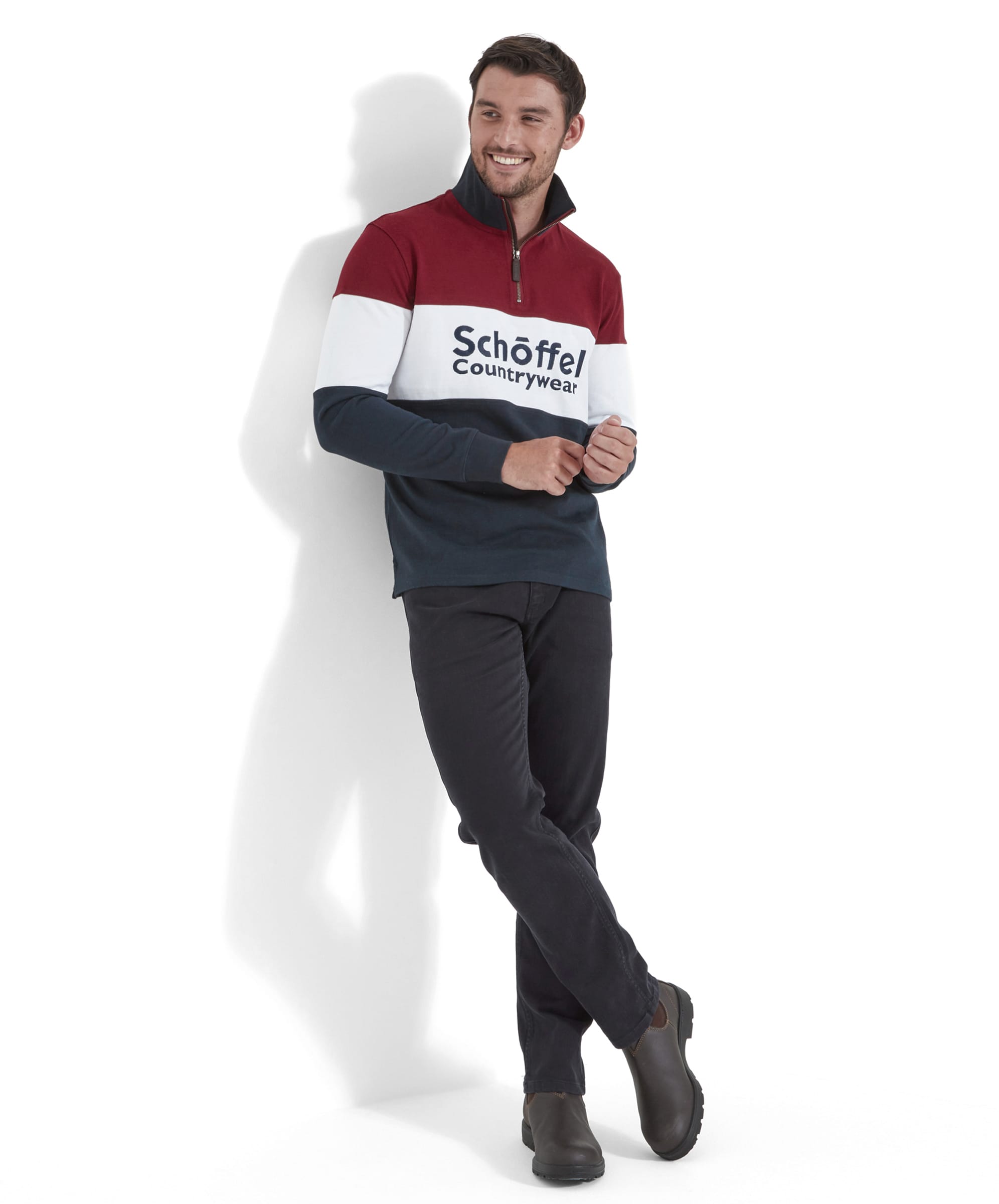 The second image displays the man standing against a white wall, smiling with his arms slightly bent and hands clasped, wearing the Schöffel Exeter Heritage 1/4 Zip for Unisex in Red, black pants, and brown shoes.