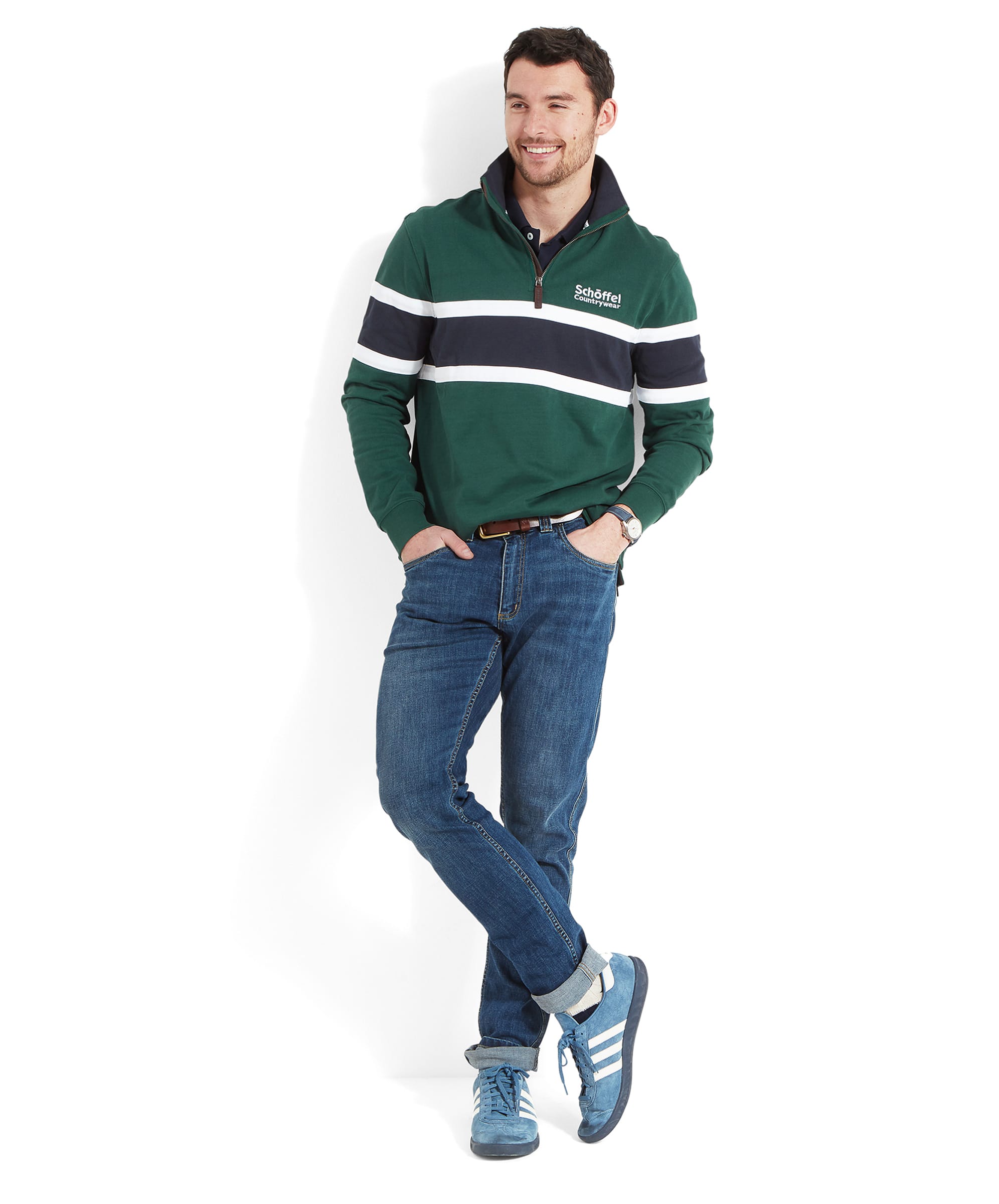 The man stands casually with his hands in his pockets, showcasing the Schöffel Exmouth Heritage 1/4 Zip for Unisex in Green, blue jeans, and blue trainers.