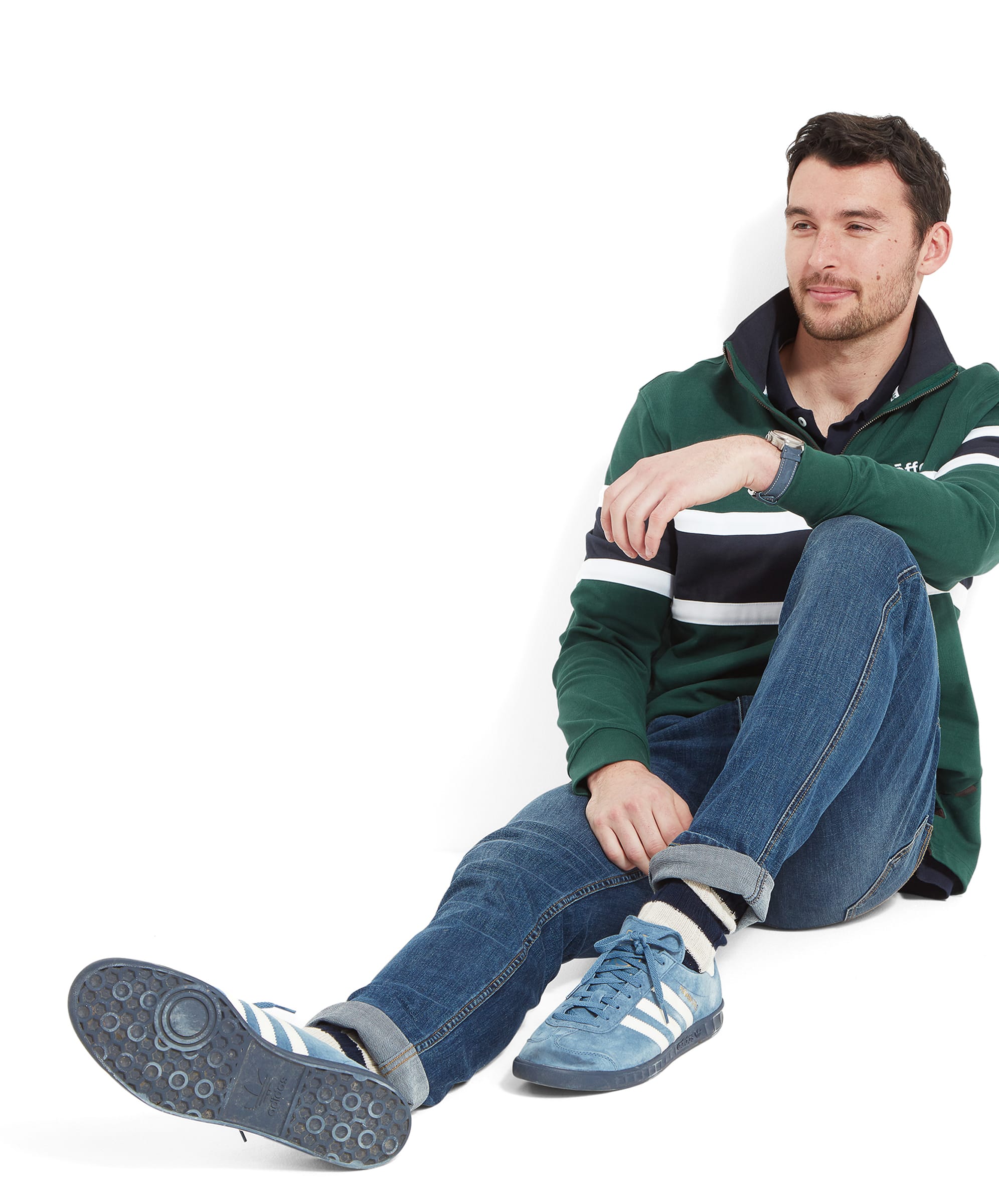 The man is sitting on the floor with his legs crossed, wearing the Schöffel Exmouth Heritage 1/4 Zip for Unisex in Green, blue jeans, and blue trainers, looking relaxed.